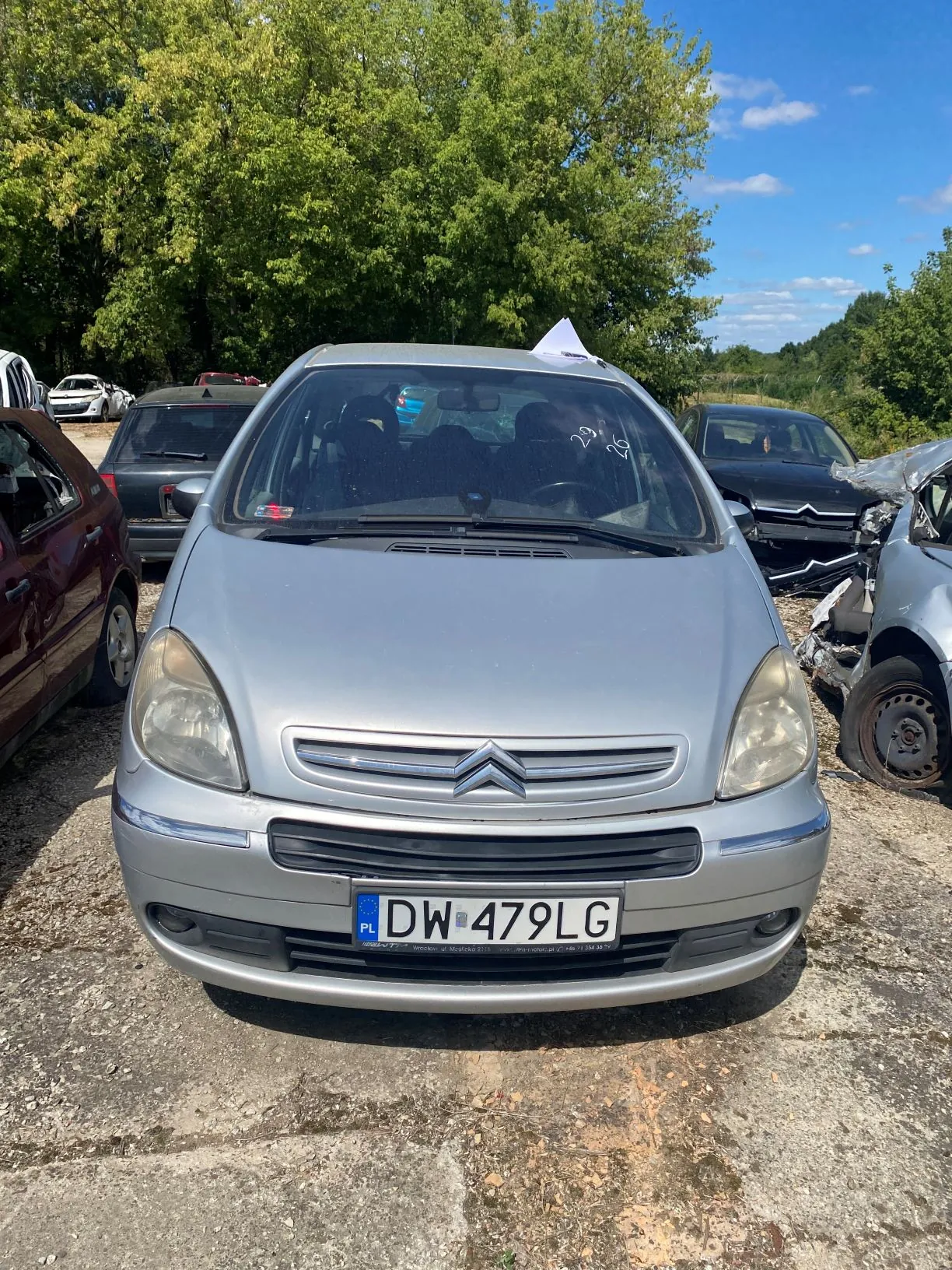 Srebrny samochód marki Citroën z tablicą rejestracyjną DW 479LG, zaparkowany na betonowym placu. Obraz jest obrócony o 90 stopni w lewo, przez co samochód znajduje się w pozycji poziomej. W tle widoczne są inne uszkodzone pojazdy oraz zielone drzewa pod niebieskim niebem.