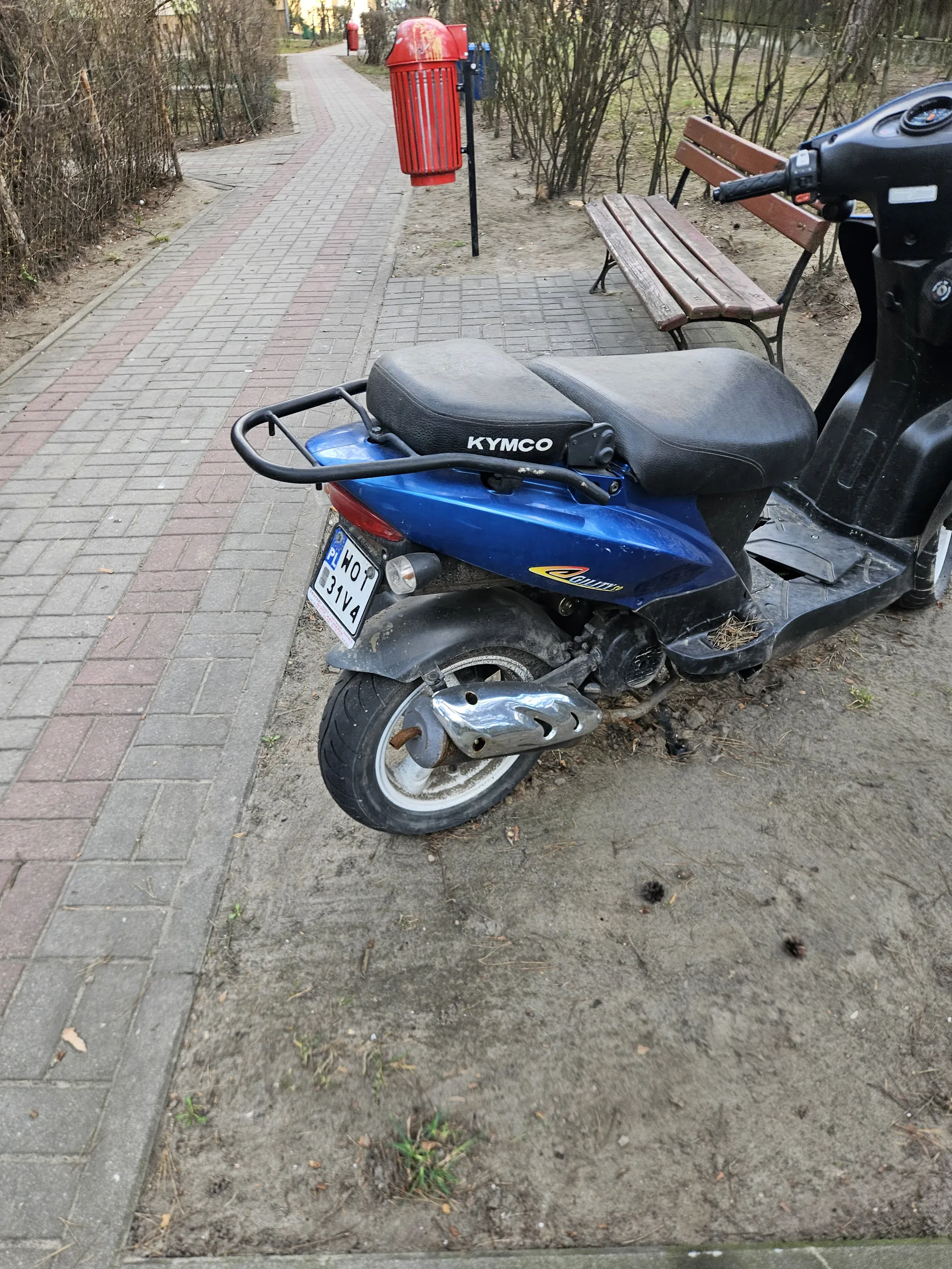 Niebieski skuter marki KYMCO z czarnym siedzeniem i tablicą rejestracyjną H01 31V4 stoi na chodniku z ceglanym wykończeniem. W tle widoczna ławka, czerwony pojemnik na śmieci oraz drzewa bez liści. Skuter znajduje się na bruku, obok niego ziemia z suchą trawą i igłami sosnowymi.