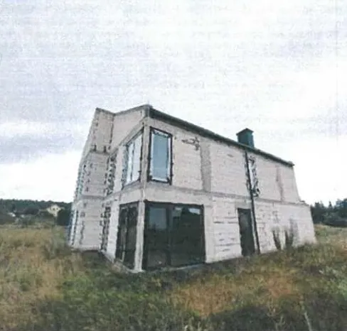 Dwupiętrowy budynek z jasnych cegieł z dużymi oknami na obu kondygnacjach, czarnymi drzwiami i kominem na dachu. Budynek znajduje się na trawiastym terenie z zaroślami, na tle zalesionego obszaru i pochmurnego nieba. Brak widocznego tekstu.