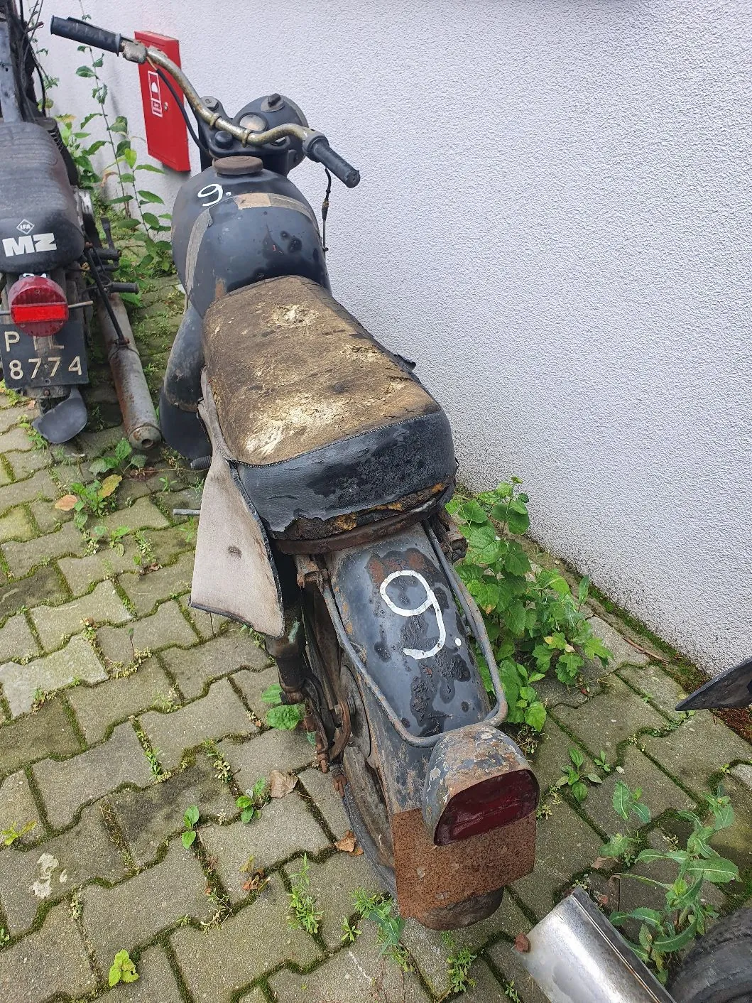 Stary, zniszczony motocykl w kolorze ciemnoszarym, zaparkowany na kostce brukowej przy białej ścianie. Pojazd ma zniszczone, brudne siedzenie, widoczne ślady rdzy na błotniku i ramie. Na baku oraz tylnym błotniku widnieje biała cyfra „9.”. W tle po lewej stronie widoczny jest fragment innego motocykla marki MZ z tablicą rejestracyjną „PL 8774”.