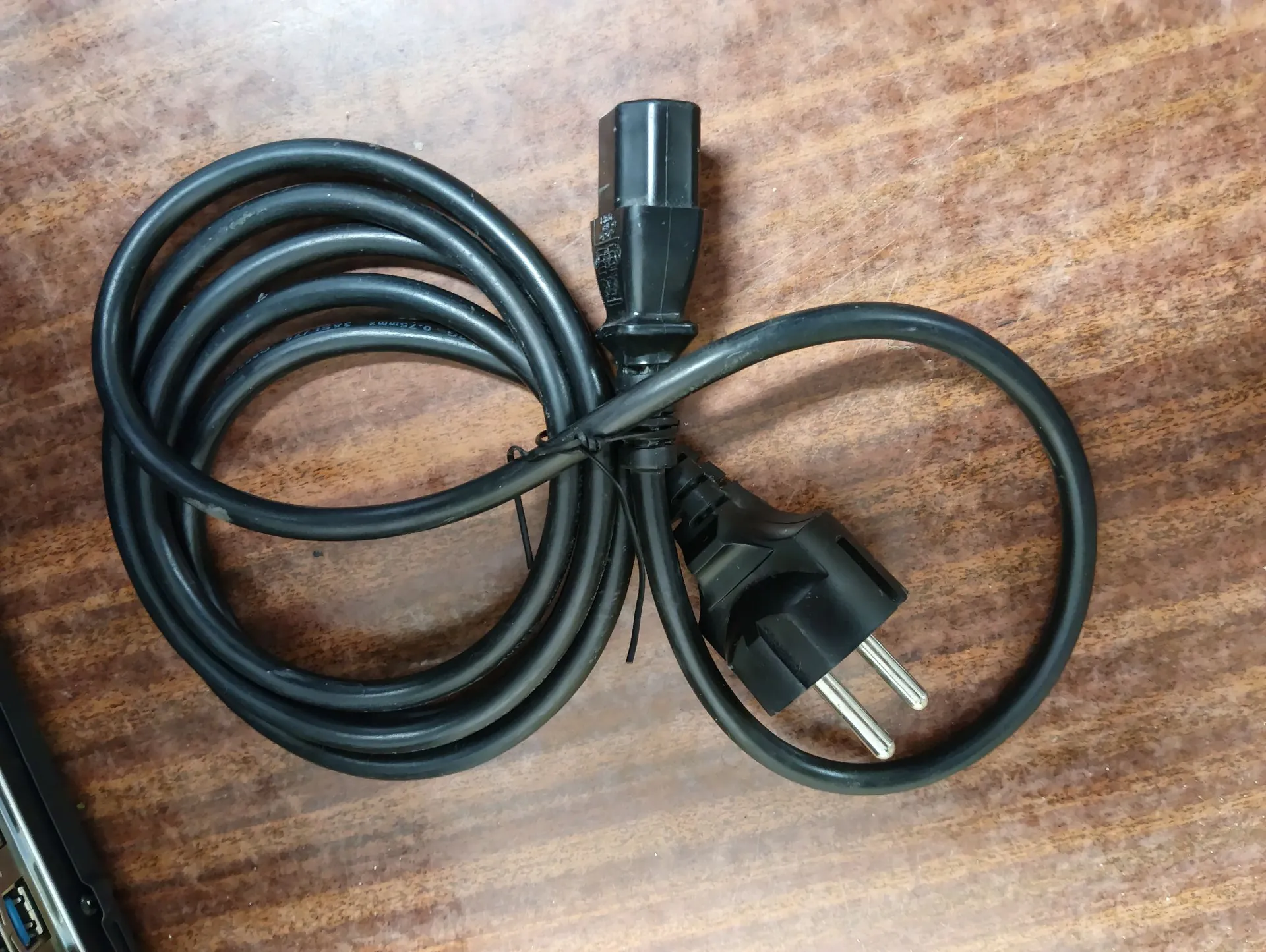 Czarny kabel zasilający z wtyczką typu C (dwie okrągłe bolce) oraz wtyczką typu C13, ułożony na brązowej, drewnianej powierzchni. Kabel jest częściowo zwinięty w pętlę.