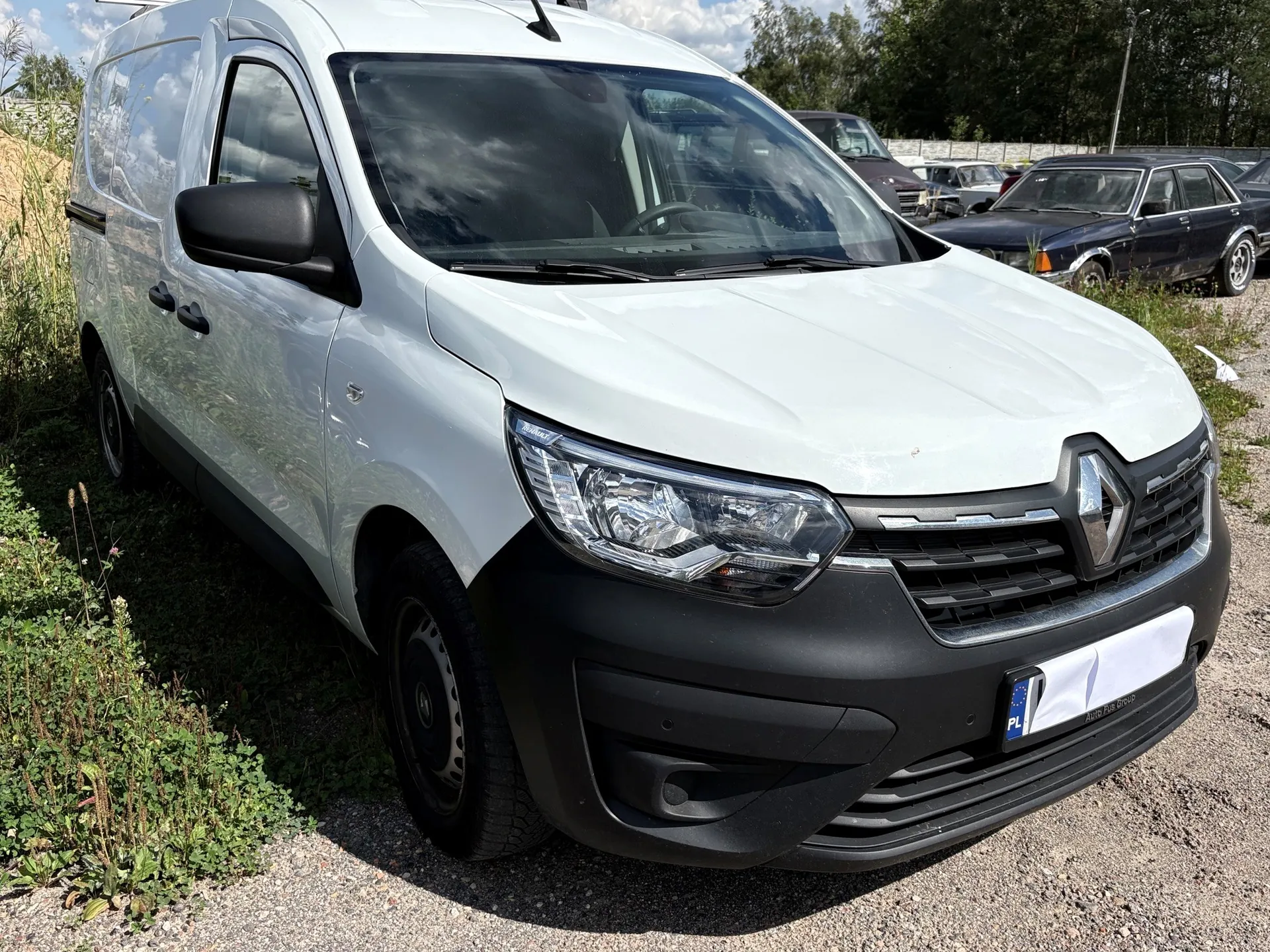 Biały samochód dostawczy marki Renault zaparkowany na żwirowym placu. Pojazd jest widoczny z przodu i z boku. W tle widoczne są inne samochody oraz pas zieleni z drzewami.