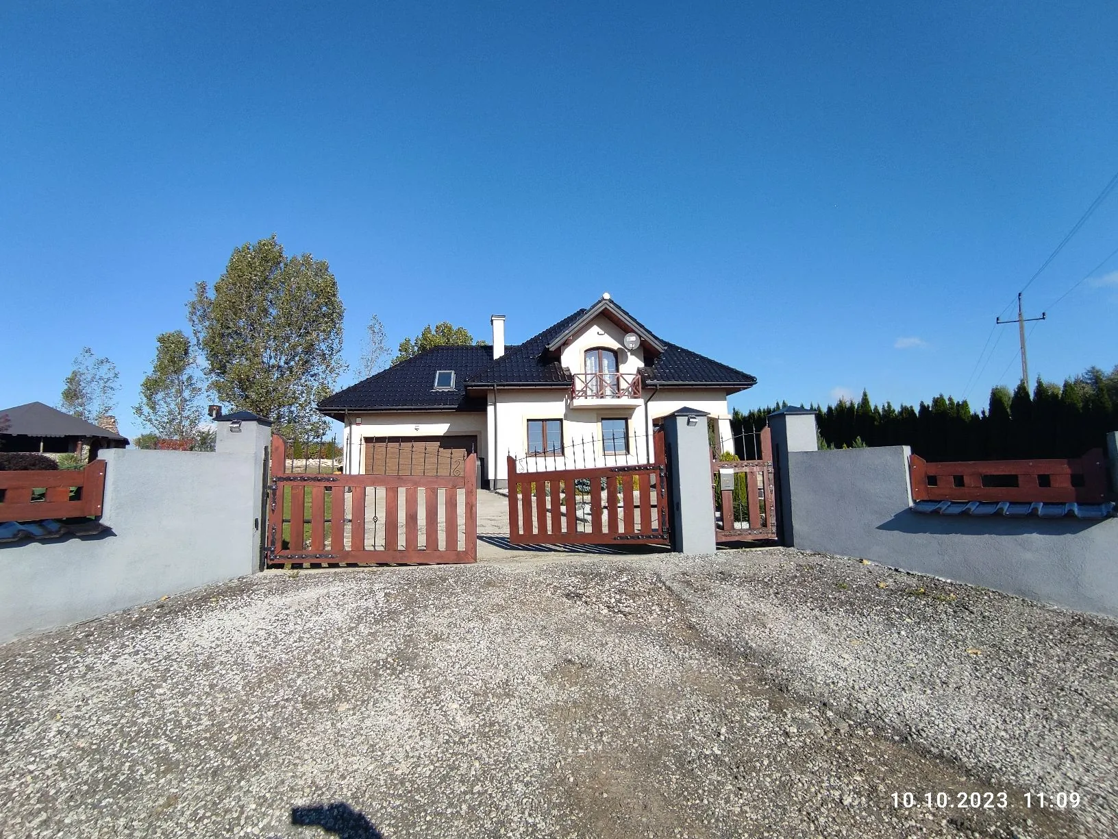 Zdjęcie jednorodzinnego domu z jasną elewacją i ciemnym, spadzistym dachem. Dom posiada balkon na piętrze oraz garaż. Przed posesją znajduje się szary betonowy mur z brązowymi, drewnianymi elementami ogrodzenia i bramą wjazdową. Przed domem widać żwirowy podjazd. W tle po lewej stronie rosną drzewa, a po prawej gęsty żywopłot. Niebo jest bezchmurne i niebieskie. W prawym dolnym rogu widnieje data i godzina: 10.10.2023 11:09.