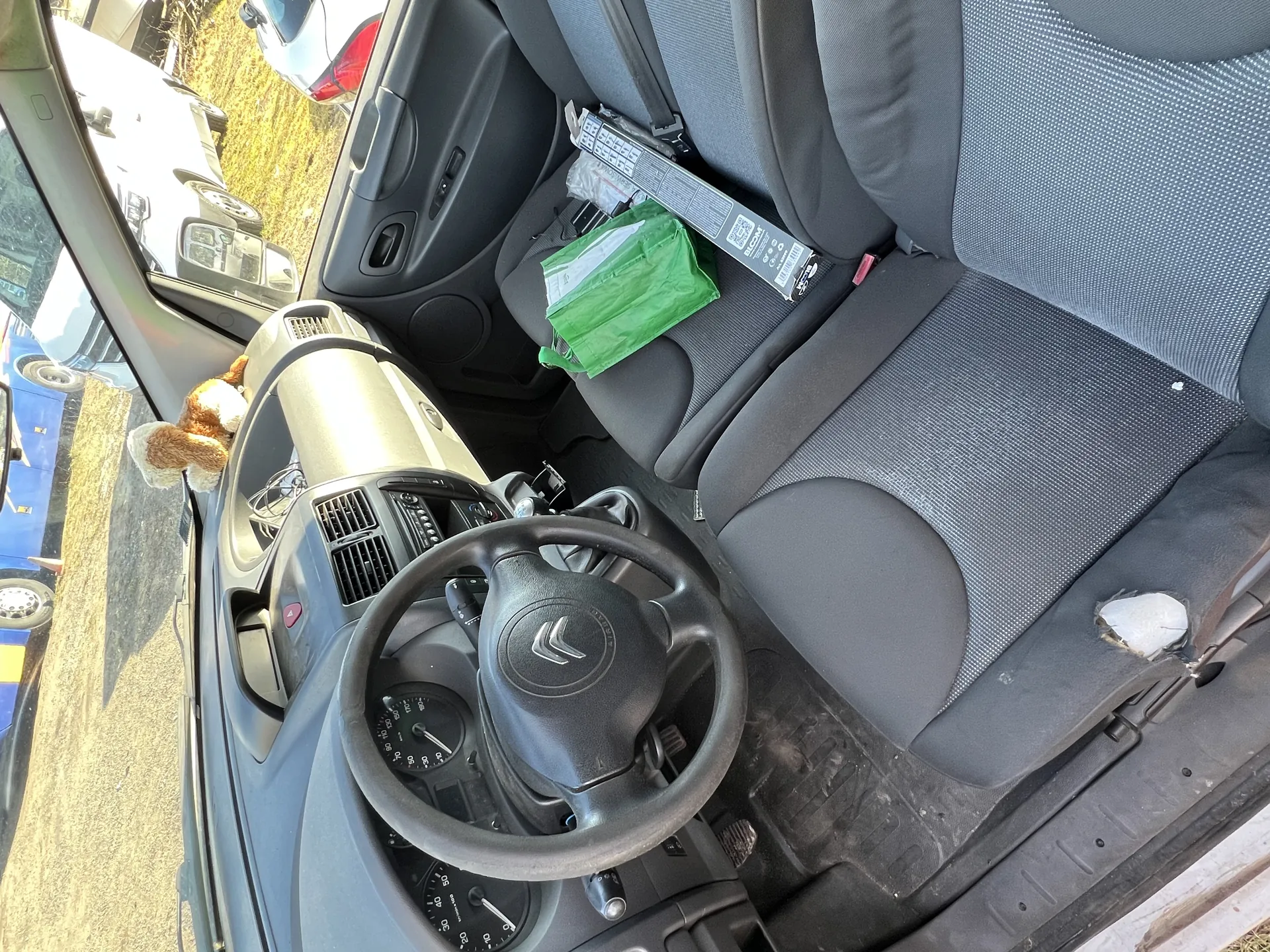 Wnętrze samochodu Citroën z widocznym kierownicą z logo marki, tablicą rozdzielczą z licznikami prędkości i obrotomierzem. Na desce rozdzielczej znajduje się pluszowy pies. Siedzenia są szare, z widocznym uszkodzeniem na siedzeniu kierowcy. Na siedzeniu pasażera leży zielona torba plastikowa oraz długie pudełko z napisem "BLOM". Poza samochodem widoczne są inne pojazdy: niebieski autobus, biały van i srebrny samochód, na tle suchego trawnika i drogi.