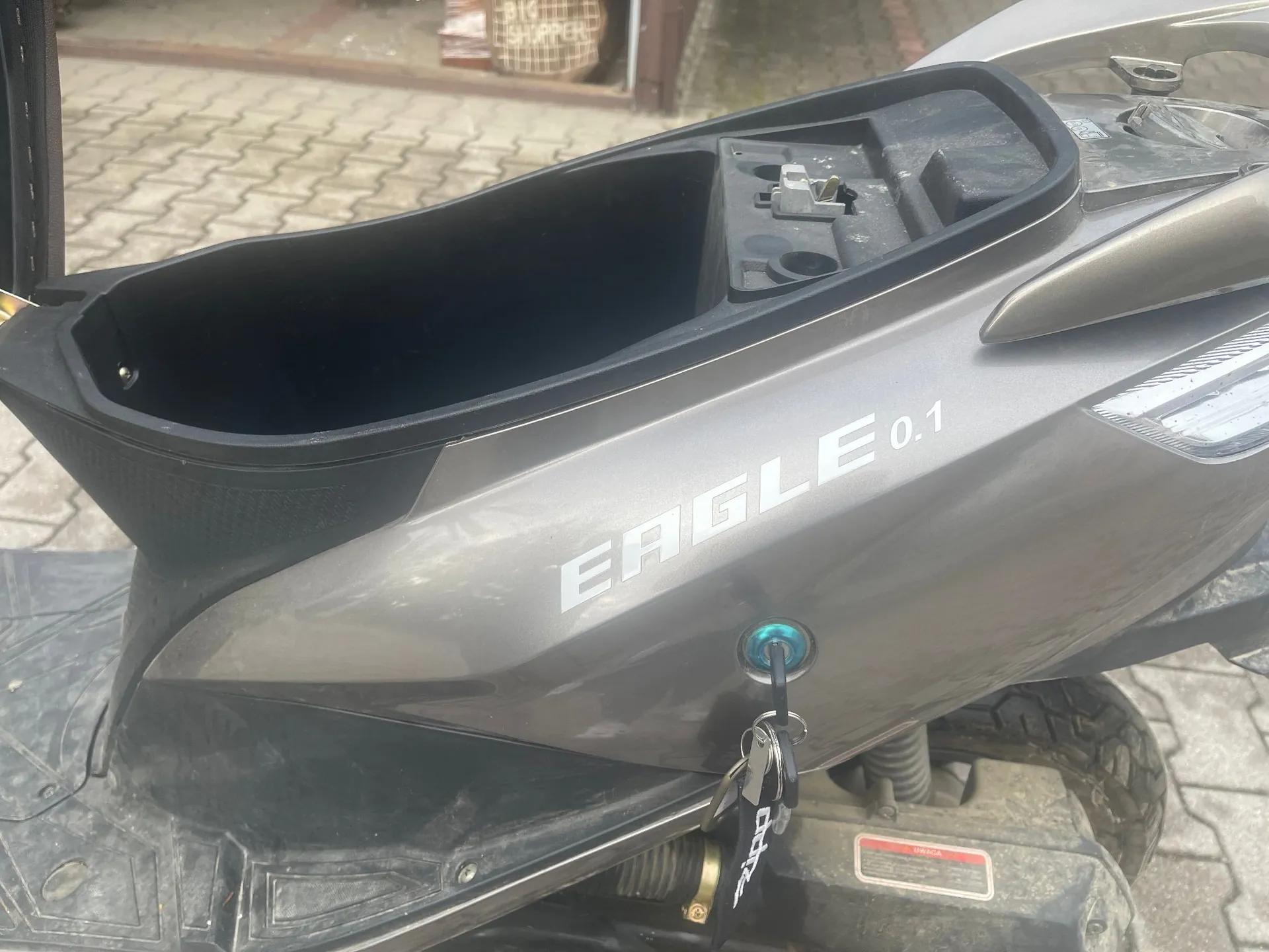 Zbliżenie na tylną część szarego skutera. Widoczny jest otwarty schowek pod siedzeniem oraz napis 'EAGLE 0.1' na bocznej obudowie. W zamku wlewu lub stacyjce znajduje się włożony kluczyk z czarnym brelokiem.