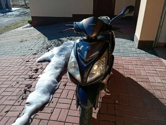 Czarny skuter marki Peugeot zaparkowany na czerwonej chodnikowej kostce. Po lewej stronie widoczna plama śniegu lub lodu. W tle budynek z brązowymi i beżowymi ścianami oraz drzwiami. Na chodniku rozrzucone są suche liście i drobne śmieci. Skuter ma włączony reflektor i widoczny logo Peugeot na przedniej części.