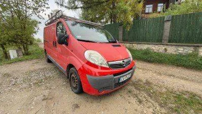 Czerwony samochód dostawczy marki Opel Vivaro zaparkowany na piaszczystej drodze. Pojazd posiada bagażnik dachowy. W tle widoczny jest zielony płot, drzewa oraz fragment budynku mieszkalnego.