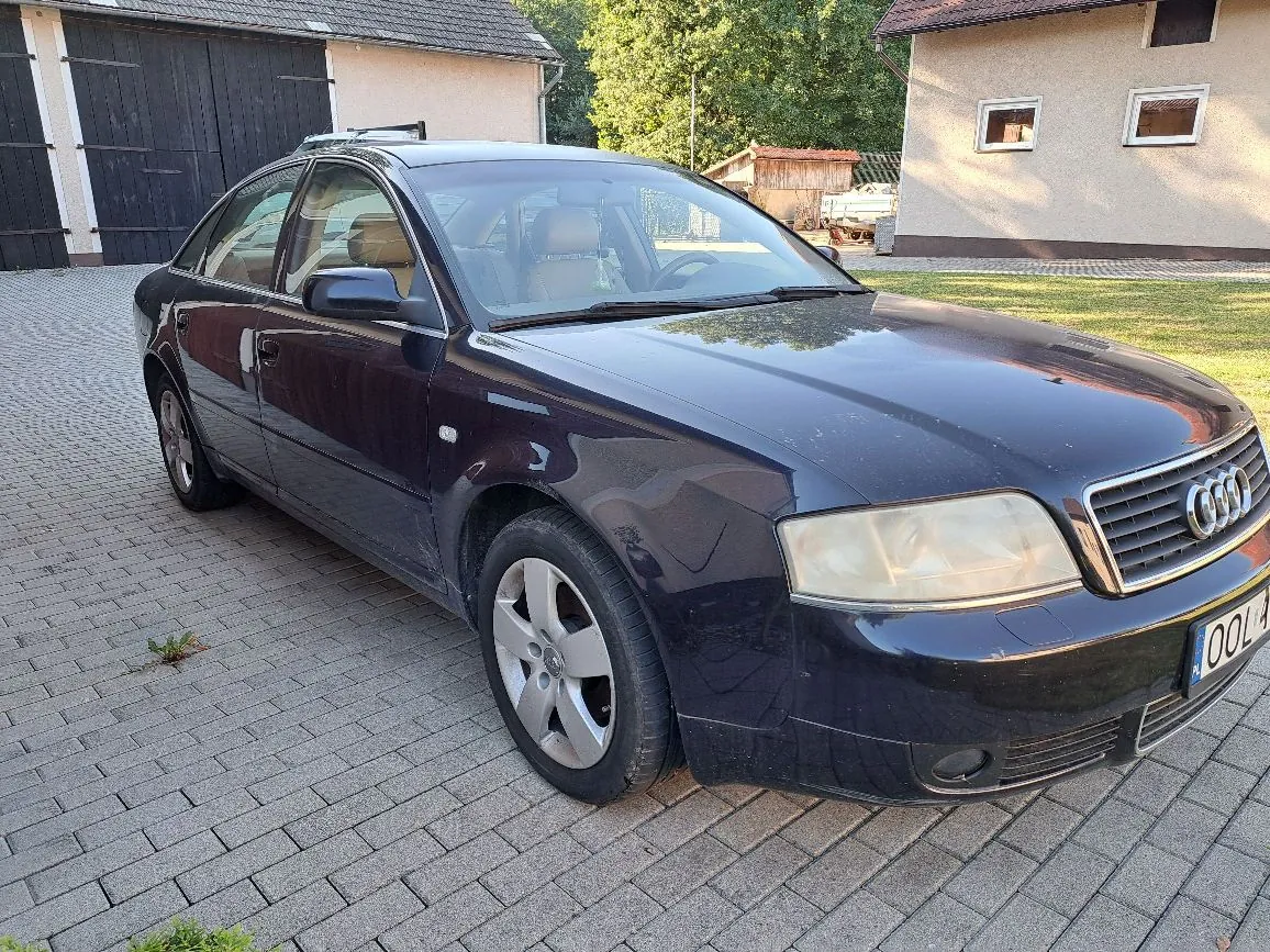 Ciemnogranatowy samochód marki Audi zaparkowany na kostce brukowej przed domem i garażem. Auto jest widoczne z przodu i z boku, ma srebrne felgi aluminiowe i widoczną część tablicy rejestracyjnej z numerem kończącym się na 'OOL'. W tle widać budynki mieszkalne oraz zieleń.