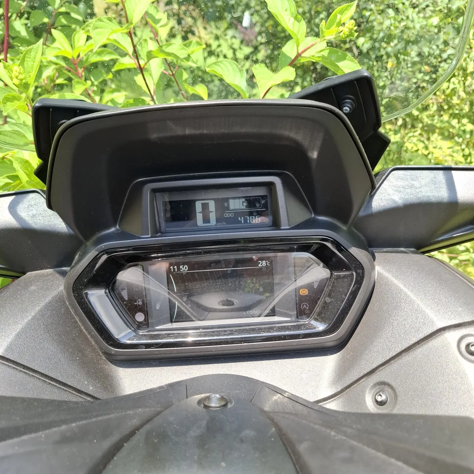 Zbliżenie na deskę rozdzielczą motocykla. Górny wyświetlacz cyfrowy pokazuje liczbę 0 oraz przebieg 4786 km. Dolny, szerszy ekran wyświetla godzinę 11:50 oraz temperaturę 28°C. Całość jest osadzona w czarnej, plastikowej obudowie, a w tle widoczne są zielone liście roślin.