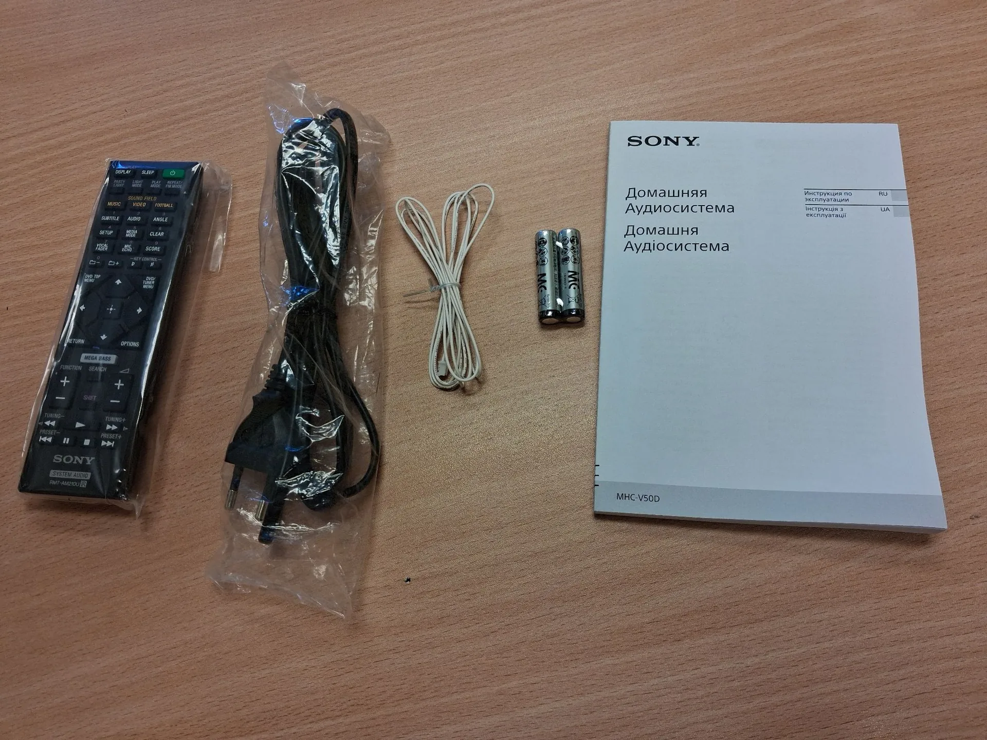 Zestaw akcesoriów do systemu audio Sony ułożony na drewnianym blacie. Od lewej znajduje się czarny pilot zdalnego sterowania z wieloma przyciskami i logo SONY na dole. Obok znajduje się czarny kabel zasilający zapakowany w przezroczystą folię, biały cienki kabel oraz dwie baterie AA. Po prawej stronie leży biała instrukcja obsługi z logo SONY i napisem w języku rosyjskim i ukraińskim: 'Домашняя Аудиосистема'. Na dole instrukcji widnieje model: MHC-V50D.