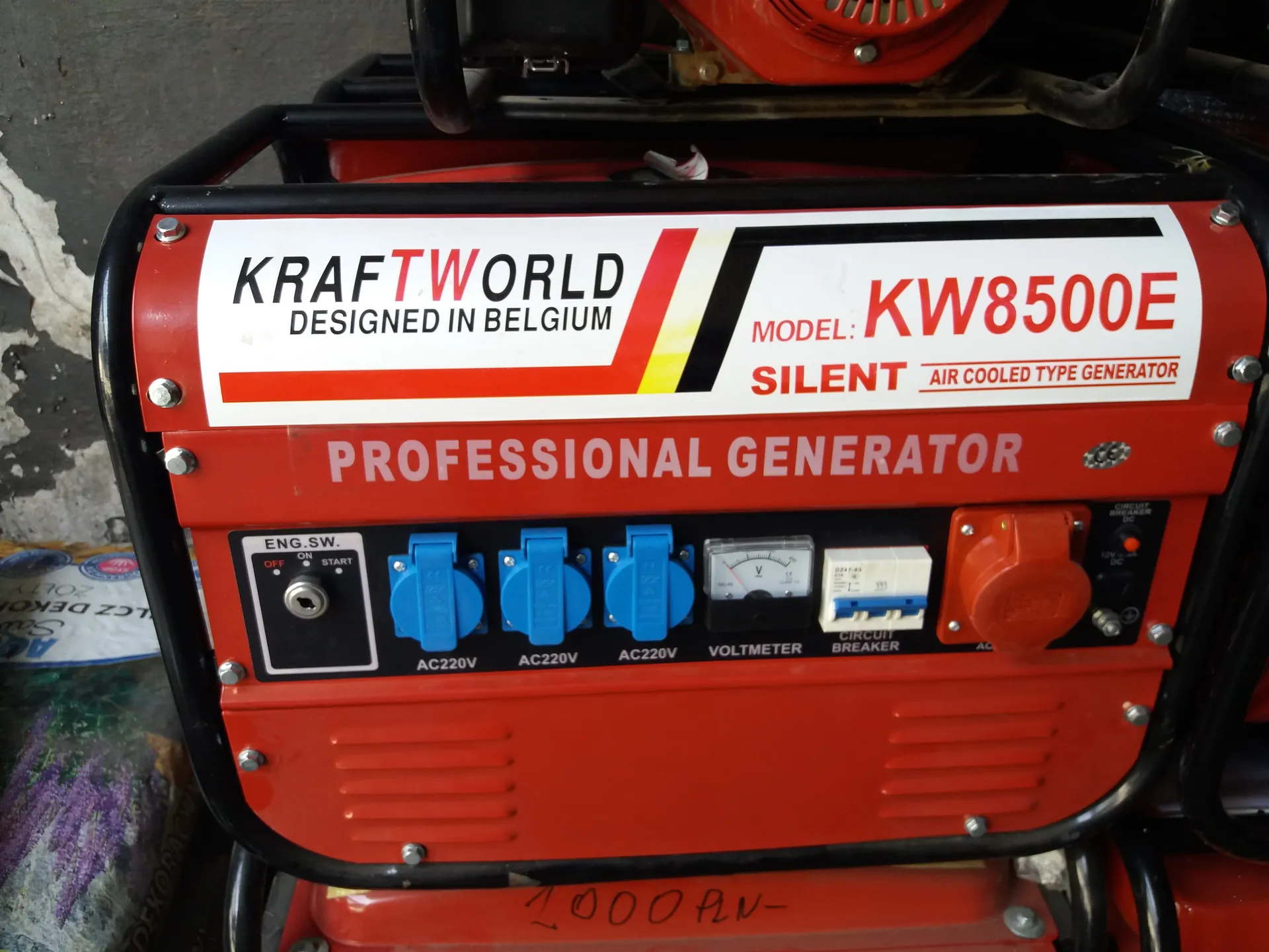 Czerwony generator prądotwórczy marki KRAFTWORLD z napisem 'DESIGNED IN BELGIUM'. Model KW8500E, typ 'SILENT AIR COOLED TYPE GENERATOR' oraz 'PROFESSIONAL GENERATOR'. Na panelu widoczne są: przełącznik 'ENG.SW.' (OFF, ON, START), trzy niebieskie gniazda 'AC220V', woltomierz, wyłącznik 'CIRCUIT BREAKER' oraz czerwony przycisk. W tle fragment betonowej ściany i worki.