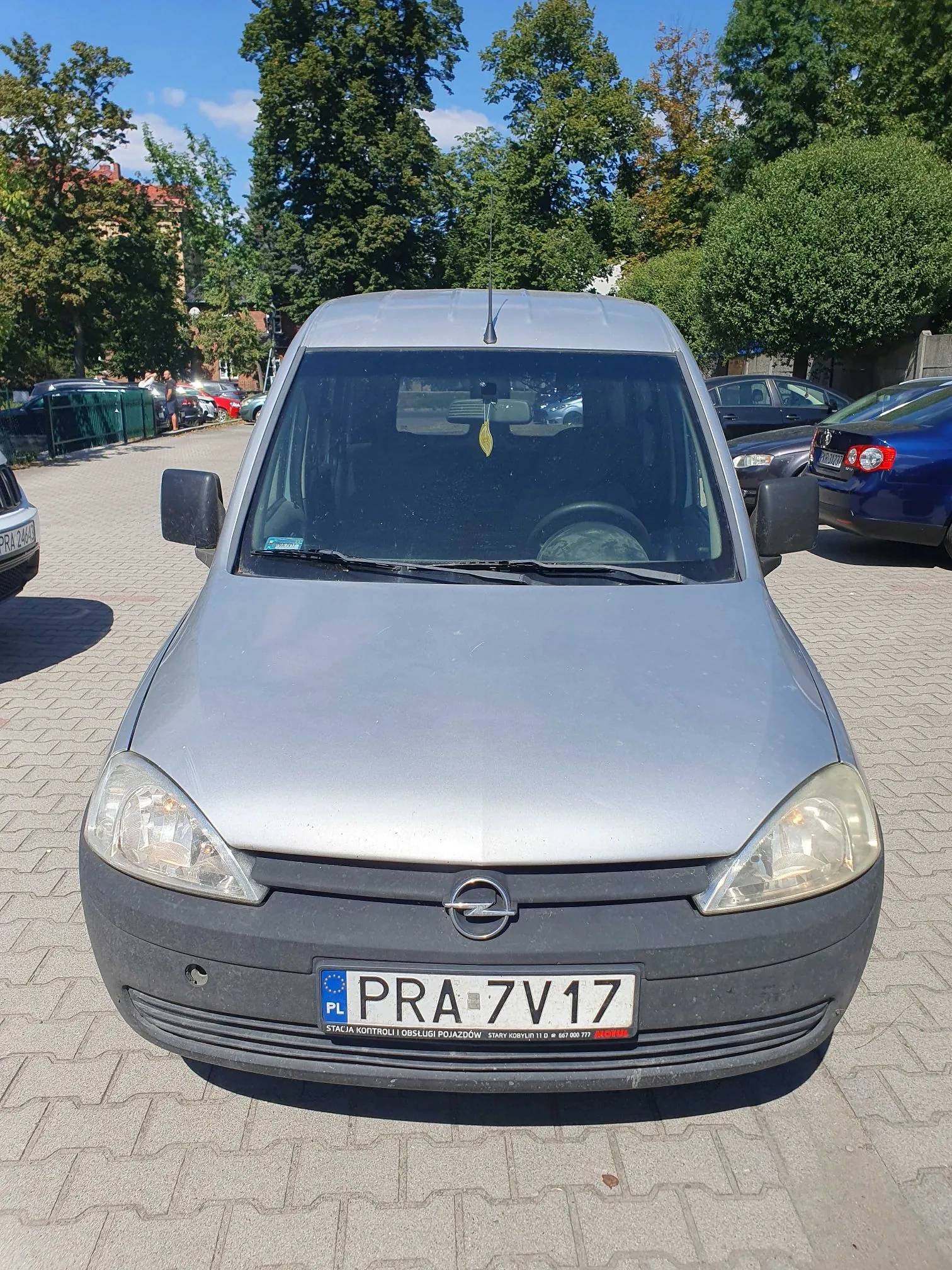 Srebrny samochód dostawczy marki Opel zaparkowany przodem do obiektywu na kostce brukowej. Pojazd posiada polską tablicę rejestracyjną o numerze PRA 7V17. Poniżej tablicy znajduje się czarny pasek z napisem: 'STACJA KONTROLI I OBSŁUGI POJAZDÓW STARY KOWALEW 11 tel. 067 888 777'. W tle widoczne są zielone drzewa, inne zaparkowane samochody oraz fragment budynku.