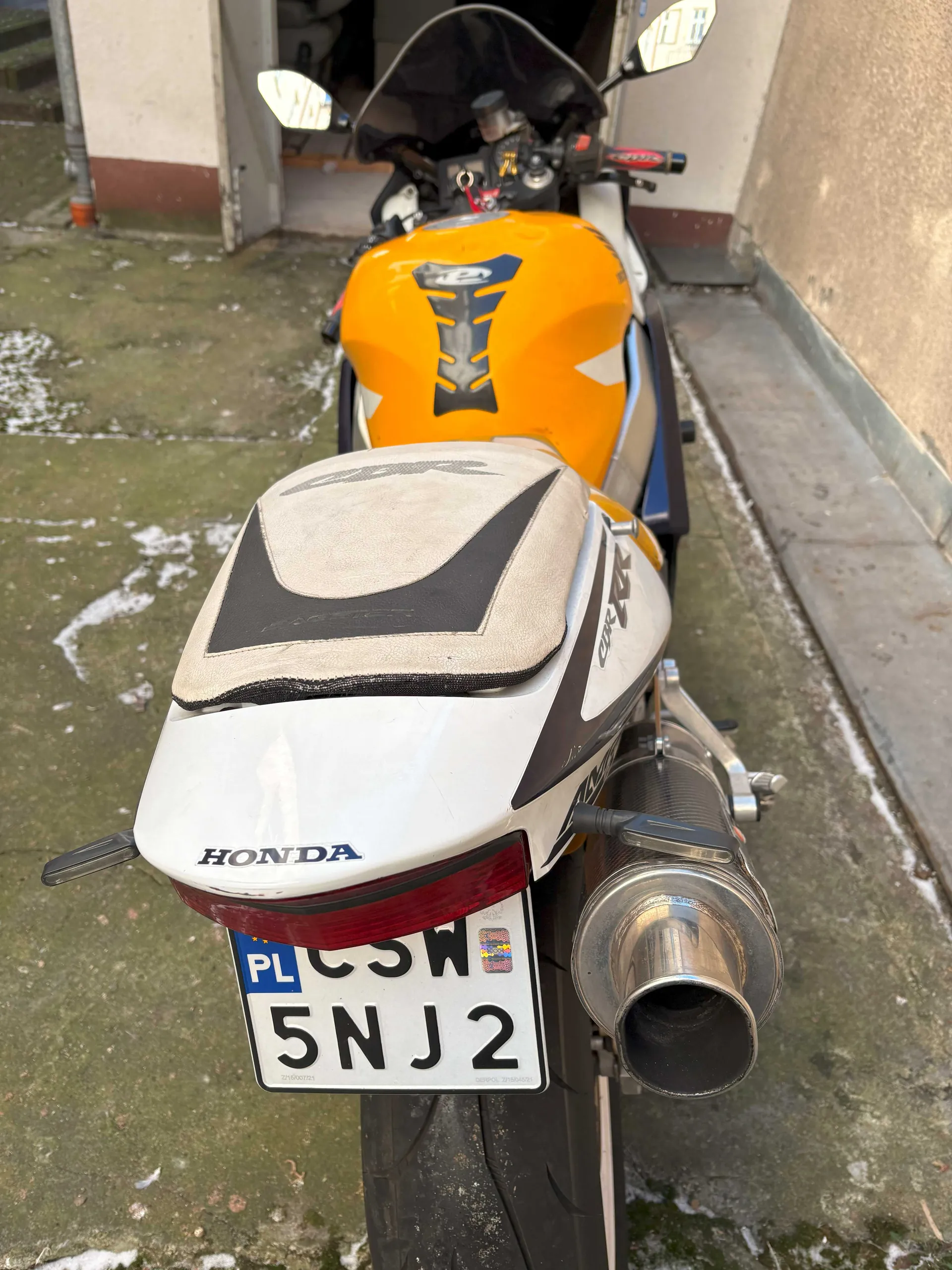 Z tyłu widoczny motocykl marki Honda w kolorze pomarańczowo-białym z czarnymi i niebieskimi akcentami. Na bagażniku widnieje napis 'HONDA', a na boku 'CBR'. Tablica rejestracyjna z polskim kodem 'PL' i numerem 'CSW 5NJ2' z kolorowym emblematem po prawej stronie. W tle betonowy podjazd z resztkami śniegu oraz budynek z otwartą bramą garażową. Widoczne są lusterka boczne, wydech i czerwone światło tylnego reflektora.