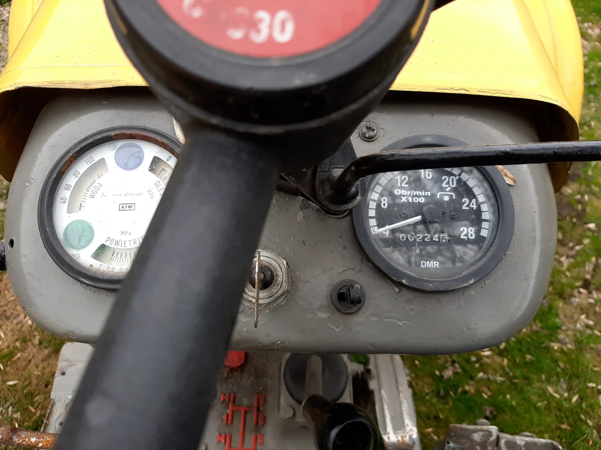Zbliżenie na panel z dwoma wskaźnikami. Po lewej stronie wskaźnik z napisami 'WODA', 'OLEJ', 'POW. IETRZ', 'KFM' oraz skalą od 0 do 100. Po prawej stronie wskaźnik z napisem 'Obr/min x100', liczbami 8, 12, 16, 20, 24, 28 i oznaczeniem 'DMR'. Na górze czerwony element z białą cyfrą '30'. Tło z zieloną trawą i częścią żółtej obudowy.