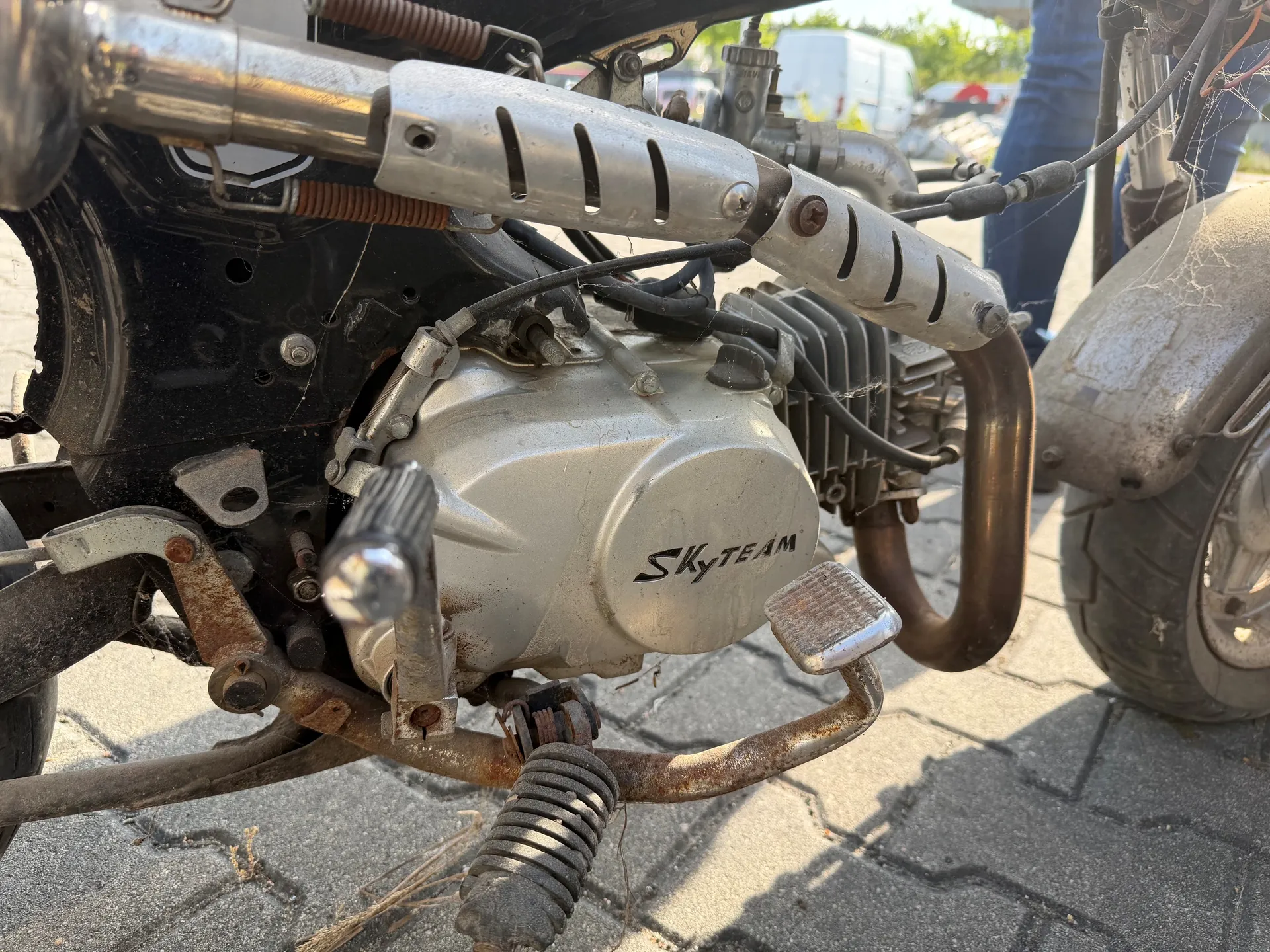 Zbliżenie na silnik motocykla marki SKyTEAM. Widoczne są metalowe rury wydechowe, rdzewiejące elementy konstrukcyjne, pedał oraz siateczka pająka na częściach silnika. W tle osoba w niebieskich dżinsach i brukowana powierzchnia.