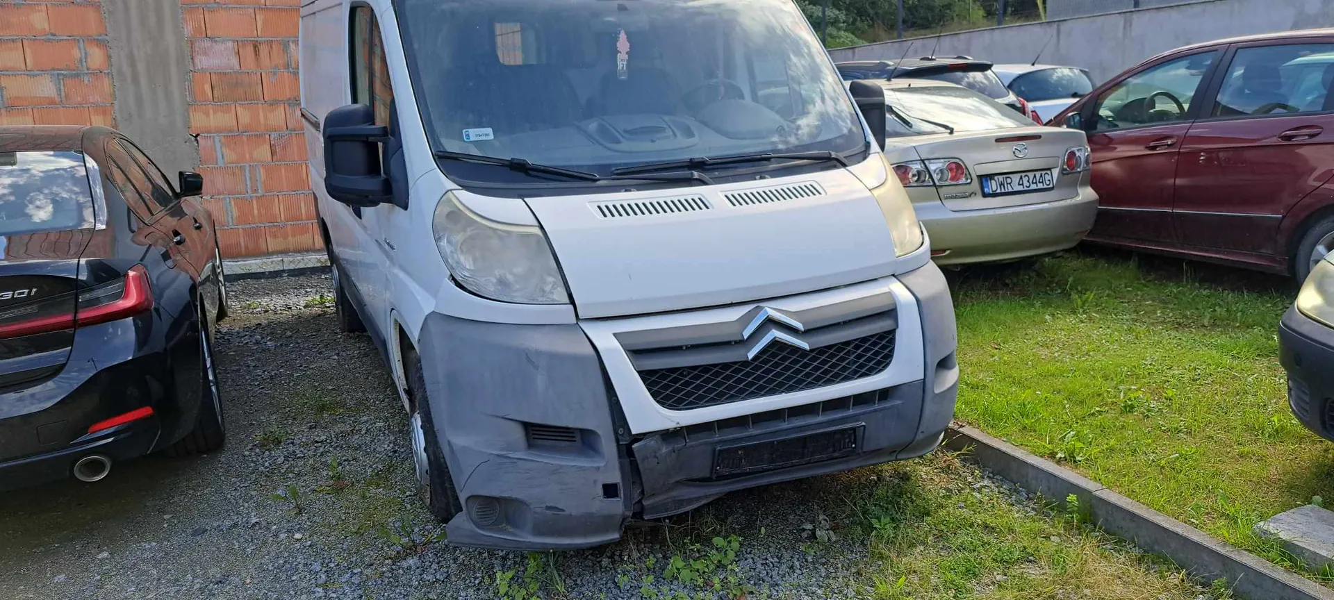 Zdjęcie przedstawiające biały samochód dostawczy marki Citroën zaparkowany na żwirowym podjeździe. Pojazd ma uszkodzony przedni zderzak po prawej stronie. Po lewej stronie widoczny jest tył czarnego samochodu osobowego, a po prawej tył białego samochodu z tablicą rejestracyjną DWR 4344G oraz fragment czerwonego samochodu. W tle znajduje się ściana z czerwonej cegły oraz zielona trawa.