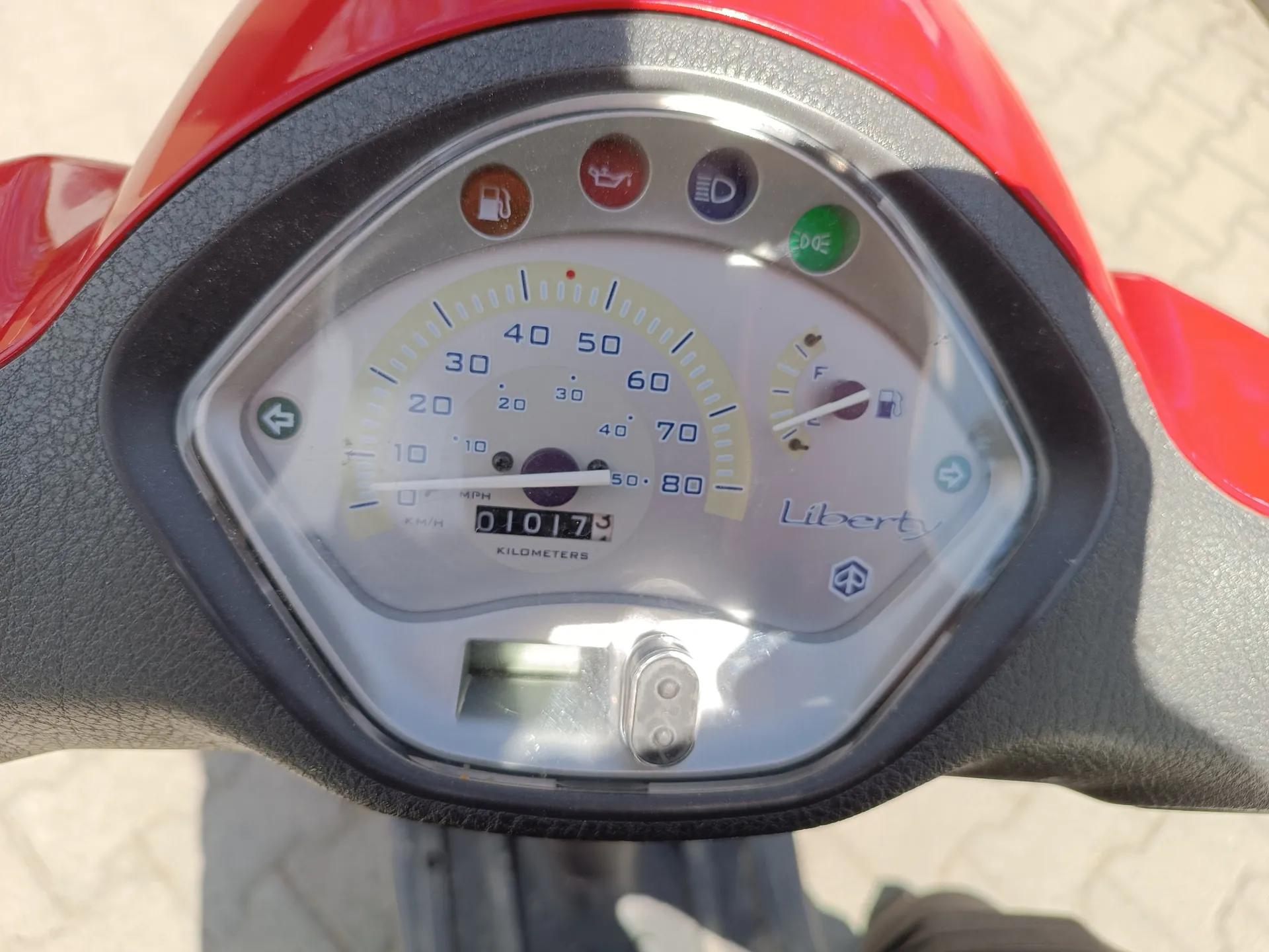 Zbliżenie na deski rozdzielcze skutera marki Liberty. Widoczny jest prędkościomierz z zakresem od 0 do 80 km/h, cyfrowy licznik przebiegu pokazujący 01017,5 kilometrów oraz wskaźniki: paliwa (pomarańczowy), ciśnienia oleju (czerwony), światła (niebieski) i trybu ECON (zielony). Po prawej stronie znajduje się wskaźnik paliwa z oznaczeniem F. Tło przedstawia brukowaną powierzchnię. Skuter ma czerwone elementy nad deska rozdzielczą.