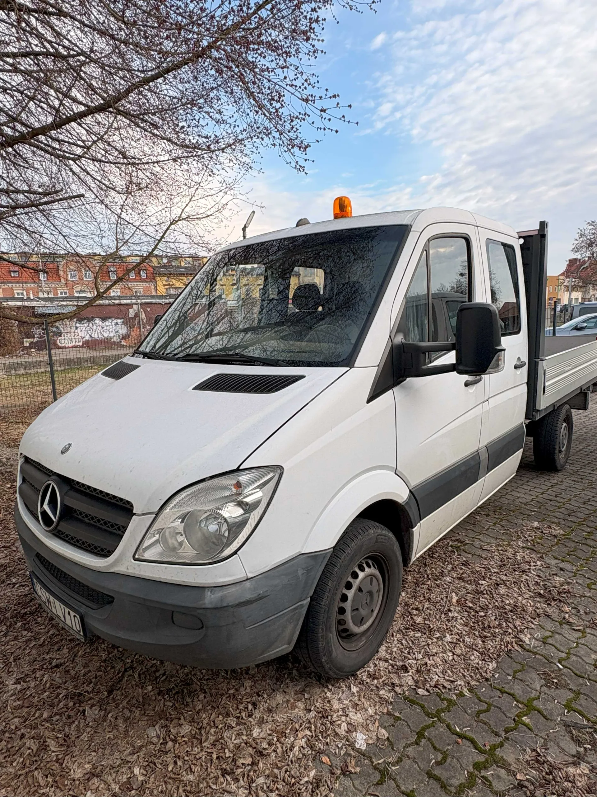 Biały samochód dostawczy marki Mercedes-Benz z czarną listwą po bokach, z widocznym logo na masce i tablicą rejestracyjną HSH V10. Na dachu pojazdu znajduje się pomarańczowe światło ostrzegawcze. Samochód ma otwarty tylny zadaszony przyczepę. W tle widoczne są budynki z czerwonymi dachami, betonowa ściana z graffiti, drzewo bez liści oraz brukowana powierzchnia pokryta suchymi liśćmi. Niebo jest częściowo zachmurzone.