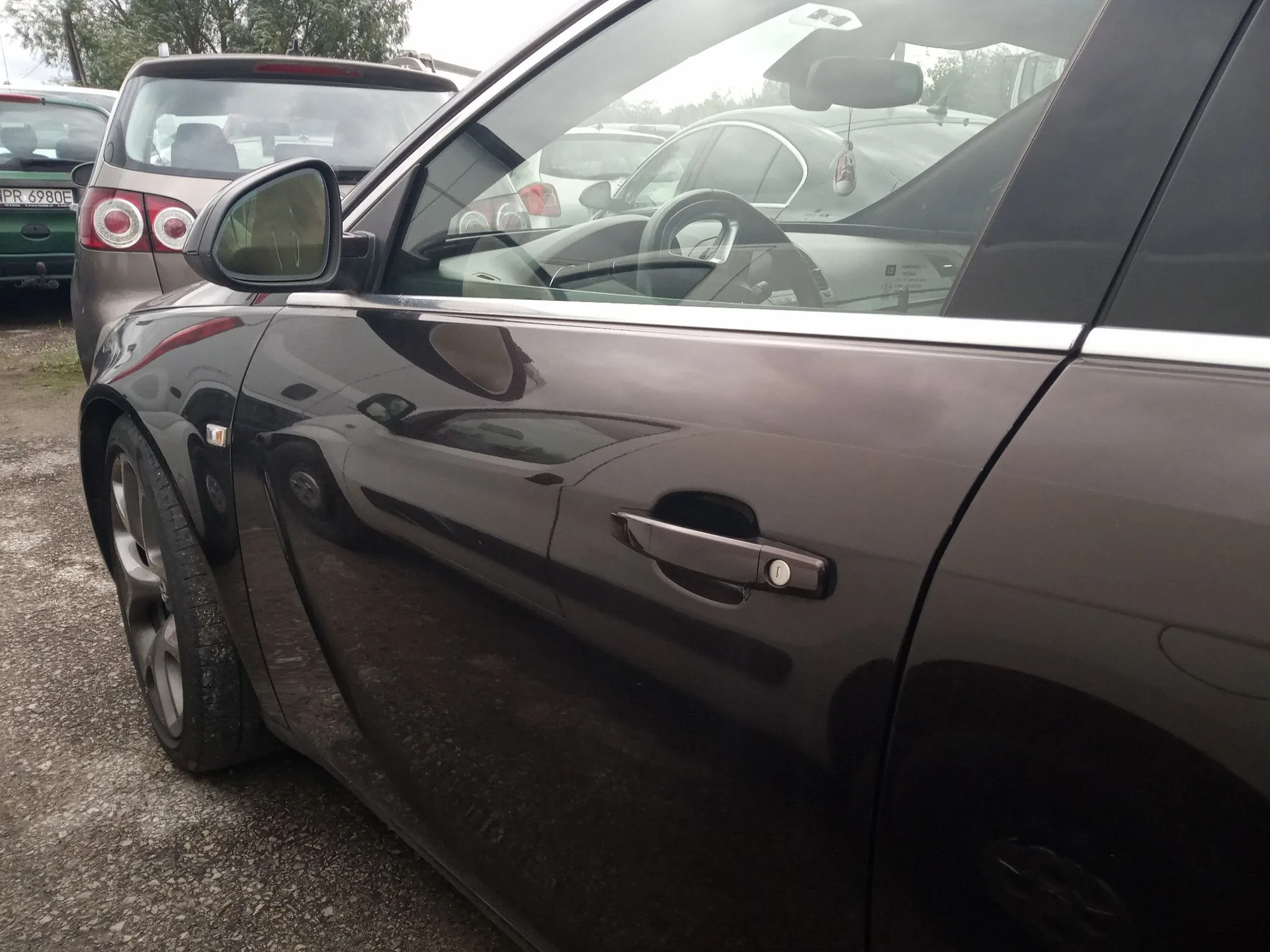 Zbliżenie na bok ciemnobrązowego samochodu, ukazujące klamkę drzwi kierowcy, lusterko boczne oraz przednie koło. W tle widoczny jest tył innego, srebrnego samochodu z tablicą rejestracyjną o numerze PR 6980E.