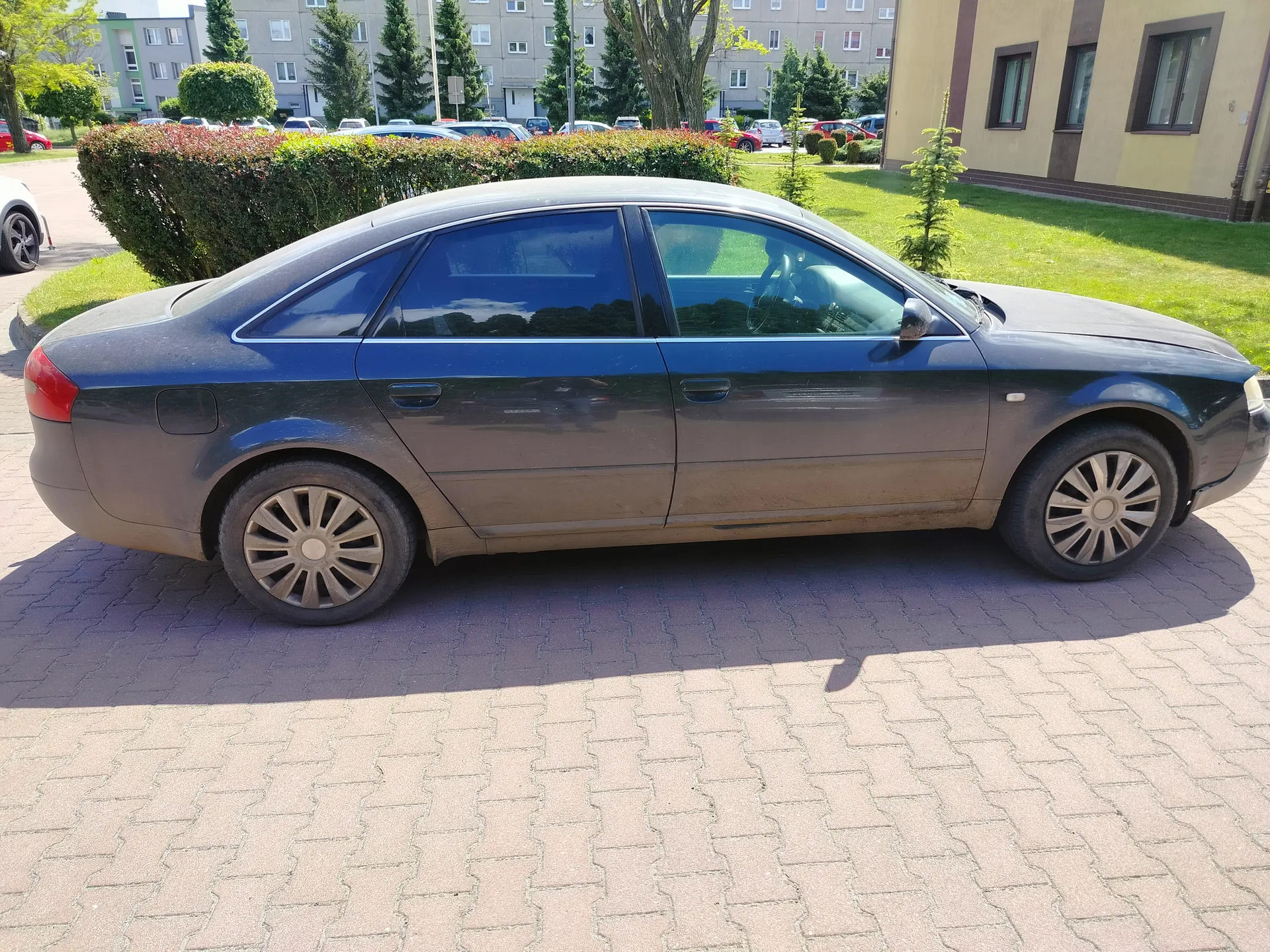 Ciemnoszary sedan zaparkowany na brukowanej powierzchni. Samochód ma brudne boki i felgi z wieloramiennym wzorem. W tle widoczne są budynki mieszkalne, zielone krzewy, drzewa oraz inne zaparkowane pojazdy. Na prawo od samochodu znajduje się żółty budynek z oknami, a w oddali szary blok mieszkalny. Trawnik i krzewy oddzielają parking od budynków.