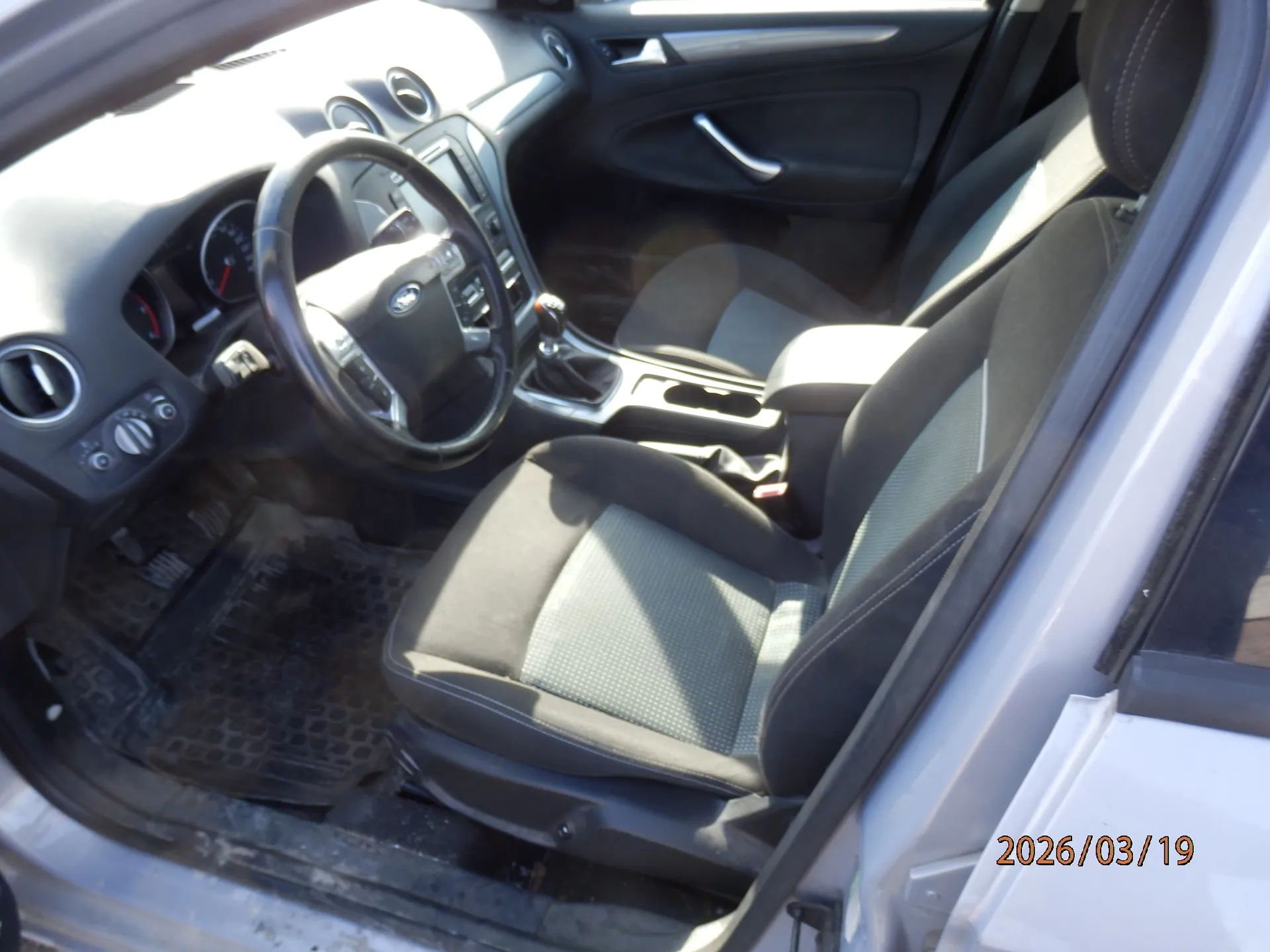 Wnętrze samochodu z widocznym kierownicą, deską rozdzielczą, siedzeniami i otwartą drzwiami po stronie kierowcy. Kierownica z logo marki, czarne siedzenia z szarymi wstawkami, deska rozdzielcza z okrągłymi wydmuchami i wskaźnikami. Na podłodze czarne gumowe dywaniki. W prawym dolnym rogu widoczny napis 2026/03/19.