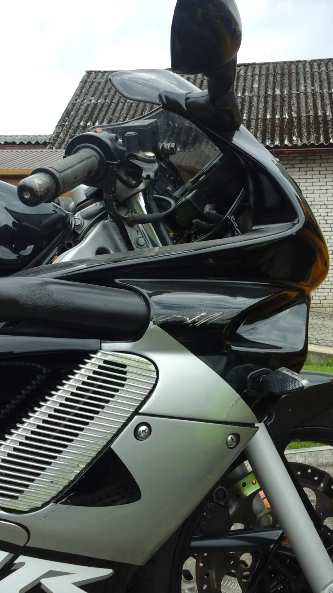 Czarno-srebrny motocykl widziany z bliska, przednia część z kierownicą, lusterkami i silnikiem. W tle budynek z dachem krytym dachówką i ceglaną ścianą oraz niebo. Na motocyklu widoczny fragment białego napisu 'M'.