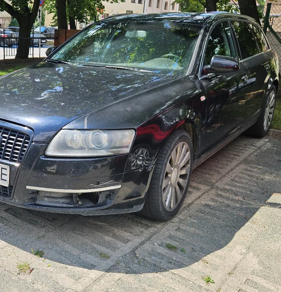Czarny samochód osobowy typu kombi zaparkowany na chodniku z kostki brukowej. Pojazd jest zakurzony, widoczne są zarysowania na przednim zderzaku po stronie pasażera. W tle widać drzewa oraz fragment ogrodzenia i innego zaparkowanego samochodu.