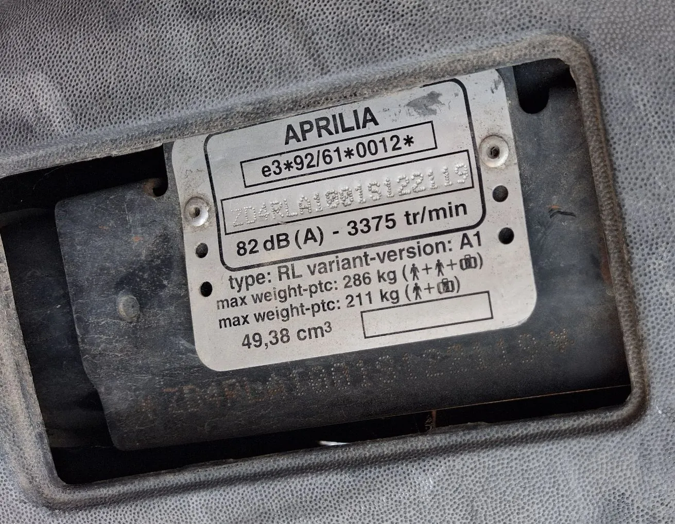 Srebrna tabliczka znamionowa marki Aprilia z czarnymi napisami. Tekst na tabliczce brzmi: 'APRILIA e3*92/61*0012*', poniżej znajduje się numer seryjny, a następnie dane techniczne: '82 dB (A) - 3375 tr/min', 'type: RL variant-version: A1', 'max weight-ptc: 286 kg (ikony dwóch osób i bagażu)', 'max weight-ptc: 211 kg (ikona jednej osoby i bagażu)', '49,38 cm3'. Tabliczka jest zamontowana w szarej, plastikowej obudowie.
