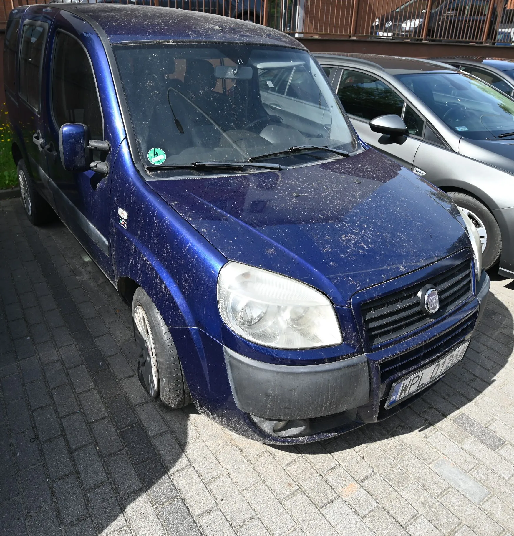 Granatowy, zabrudzony samochód dostawczy marki Fiat zaparkowany na kostce brukowej. Pojazd jest pokryty warstwą kurzu i błota. Widoczna jest tablica rejestracyjna o numerze WPL 01204 oraz naklejka w przedniej szybie.