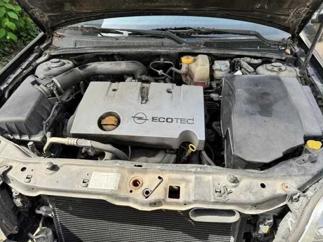 Widok z góry na komorę silnika samochodu z otwartą maską. Na środku znajduje się plastikowa osłona silnika z logo marki Opel oraz napisem 'ECOTEC'. Widoczne są elementy takie jak obudowa filtra powietrza po prawej i lewej stronie, zbiornik z płynem chłodniczym oraz chłodnica w dolnej części zdjęcia.