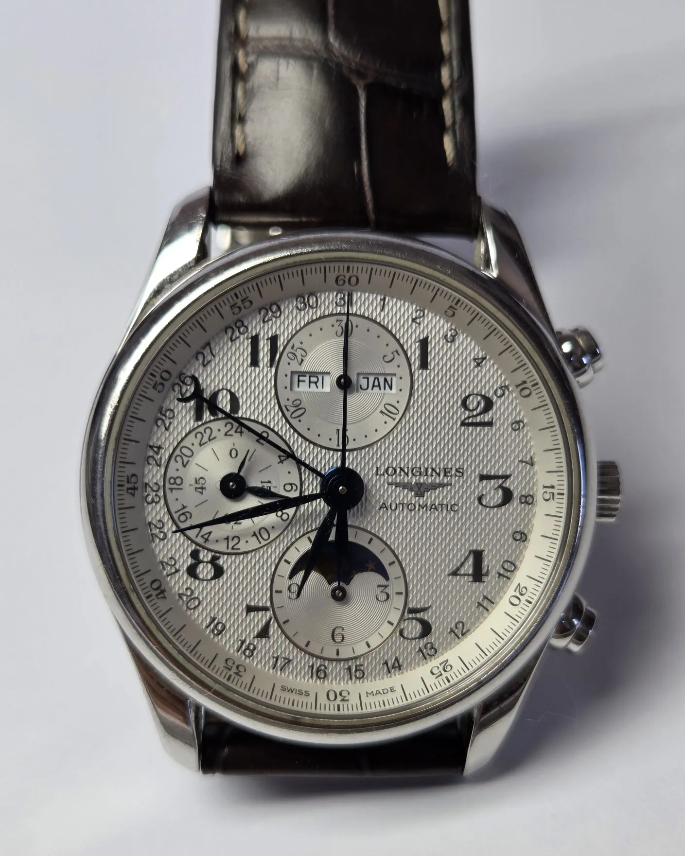 Zbliżenie na tarczę srebrnego zegarka marki Longines z automatycznym mechanizmem. Zegarek posiada białą, teksturowaną tarczę z czarnymi cyframi arabskimi oraz trzema mniejszymi tarczami pomocniczymi. Na tarczy widoczne są okienka z datą i dniem tygodnia z napisami 'FRI' oraz 'JAN'. Zegarek ma srebrną kopertę i czarny skórzany pasek.