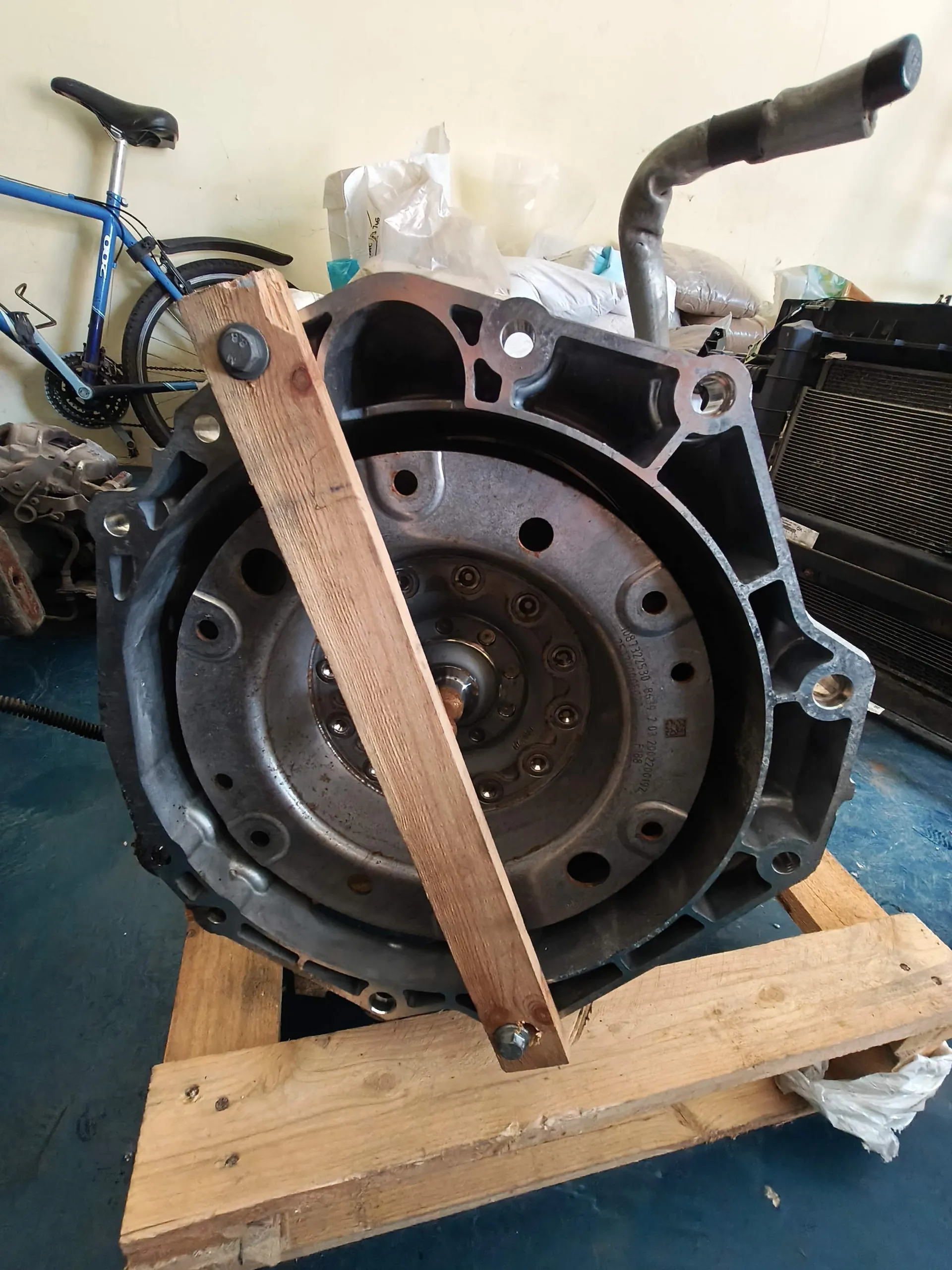 Zdjęcie przedstawiające duży, metalowy element mechaniczny, prawdopodobnie obudowę skrzyni biegów lub sprzęgła, spoczywający na drewnianej palecie. Element jest zabezpieczony drewnianą listwą przykręconą śrubami. W tle widoczny jest niebieski rower oraz inne części samochodowe i plastikowe opakowania.
