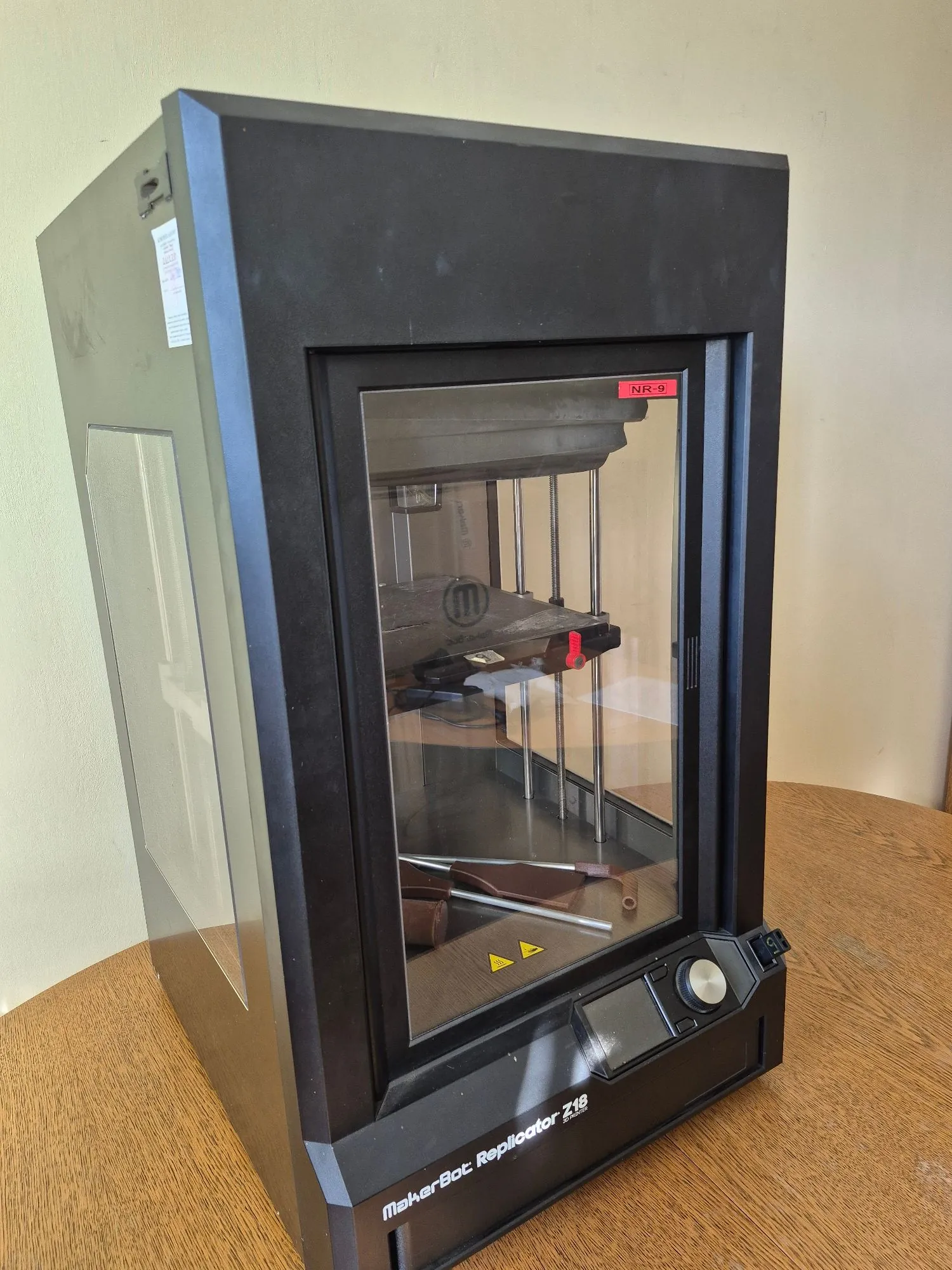 Czarna drukarka 3D MakerBot Replicator Z18 w obudowie z przezroczystą szybą z przodu i z boku. Na obudowie znajduje się czerwona naklejka z napisem 'NR-9'. Na dolnej krawce urządzenia widnieje napis 'MakerBot Replicator Z18'. Drukarka stoi na drewnianym stole na tle jasnej ściany.