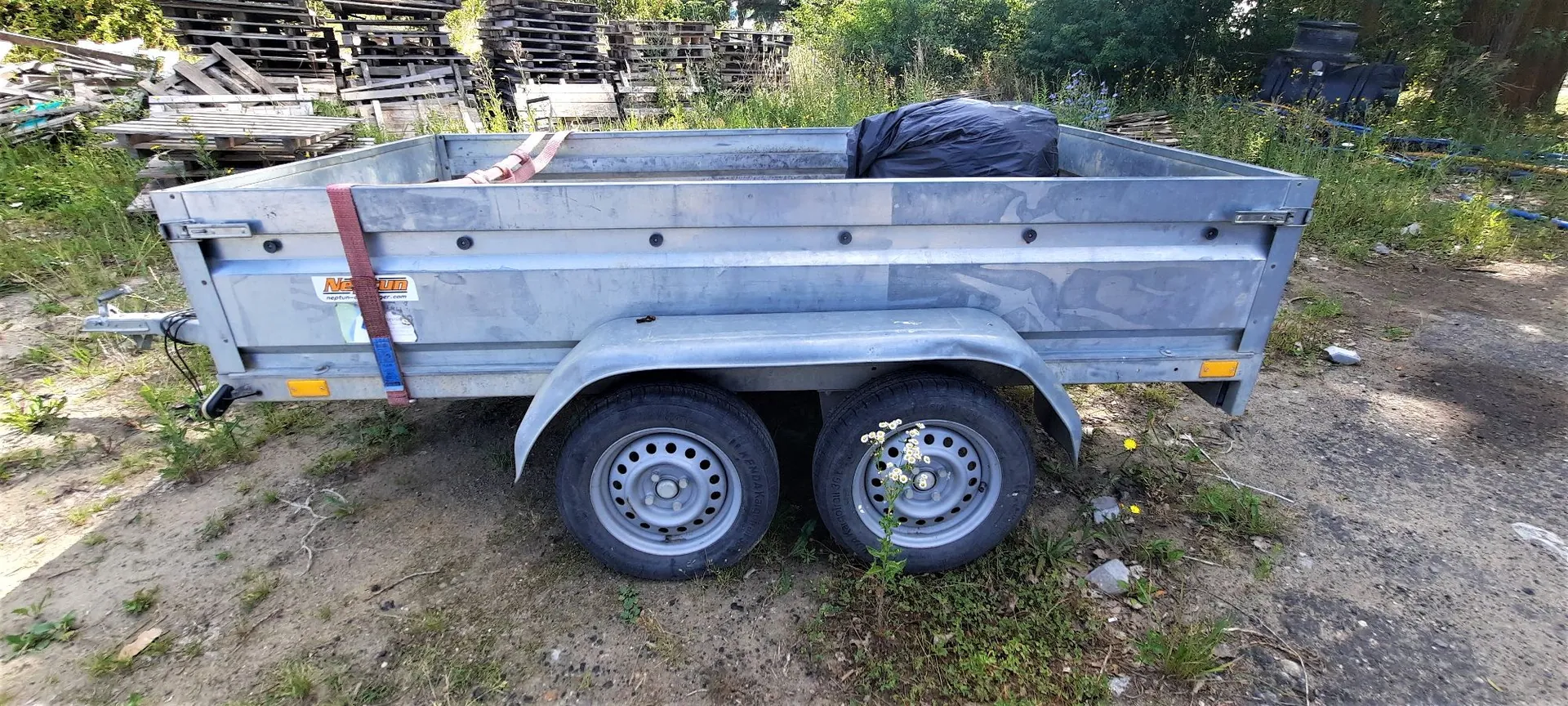 Srebrna, dwuosiowa przyczepa transportowa z ocynkowanej stali, zaparkowana na piaszczystym podłożu. Wewnątrz przyczepy znajduje się czarny worek na śmieci, a całość jest zabezpieczona czerwonym pasem transportowym. W tle widoczne są stosy drewnianych palet oraz zielona roślinność.