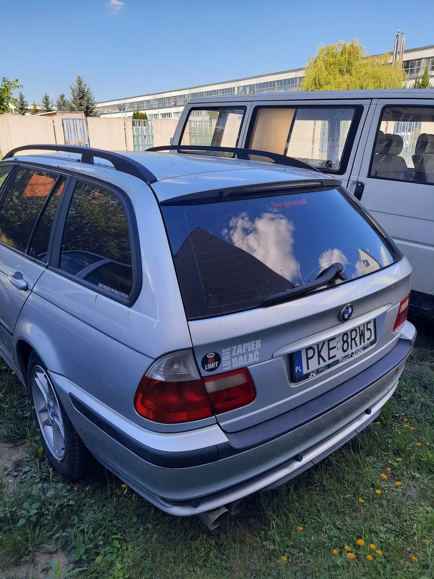 Srebrny samochód osobowy marki BMW w nadwoziu kombi, zaparkowany na trawie. Widoczny jest tył pojazdu z tablicą rejestracyjną PKE 8RW5. Na tylnej szybie znajduje się napis 'See you in hell', a na klapie bagażnika naklejki z napisami 'LIMIT' oraz 'ZAPIER DALAĆ'. W tle widoczny jest biały samochód dostawczy oraz długi, niski budynek przemysłowy pod niebieskim niebem.