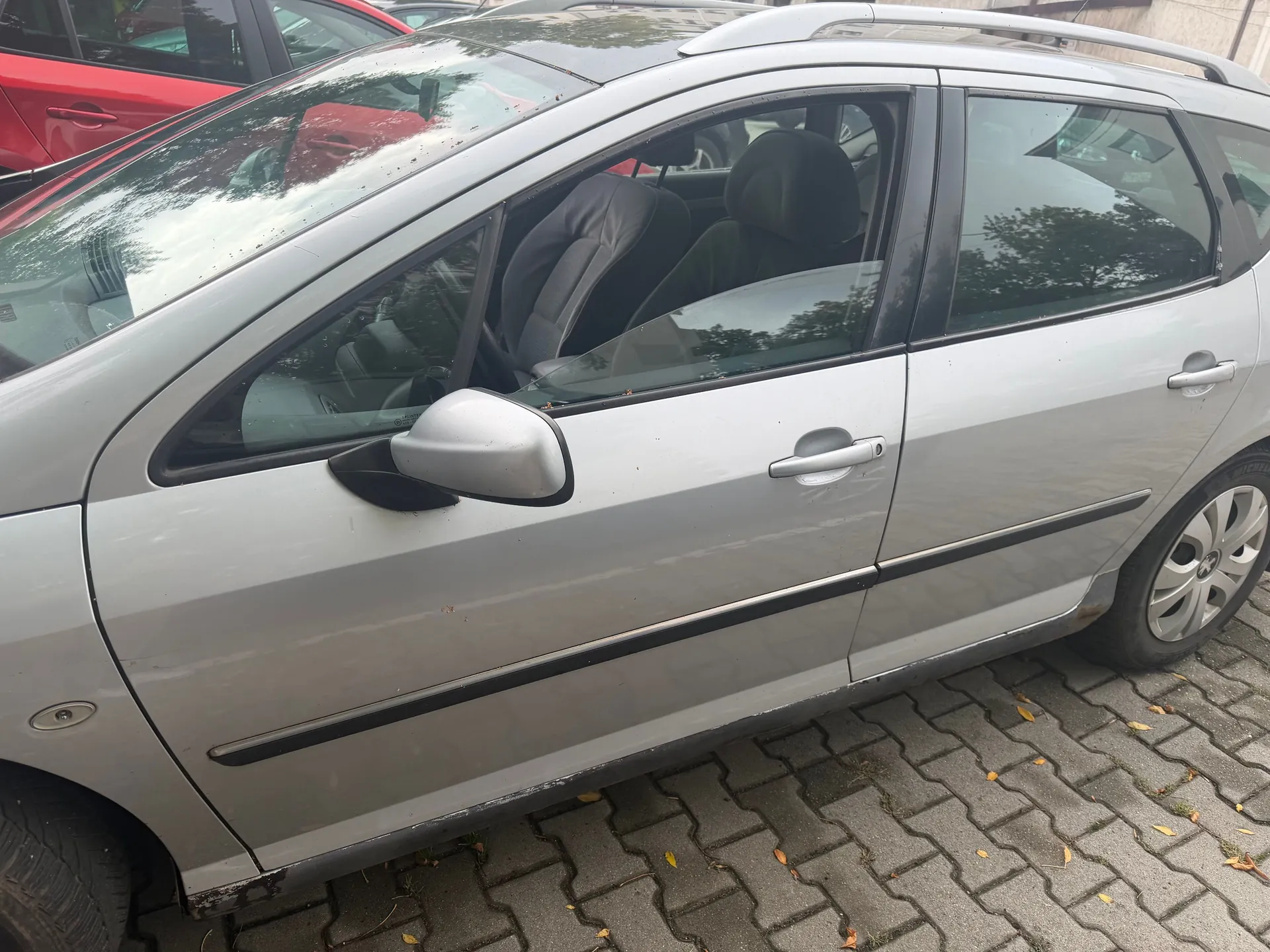 Srebrny samochód osobowy typu kombi zaparkowany na kostce brukowej. Widoczny jest bok pojazdu z przednimi i tylnymi drzwiami, lusterkiem bocznym oraz kołami. W tle widać fragment czerwonego samochodu.