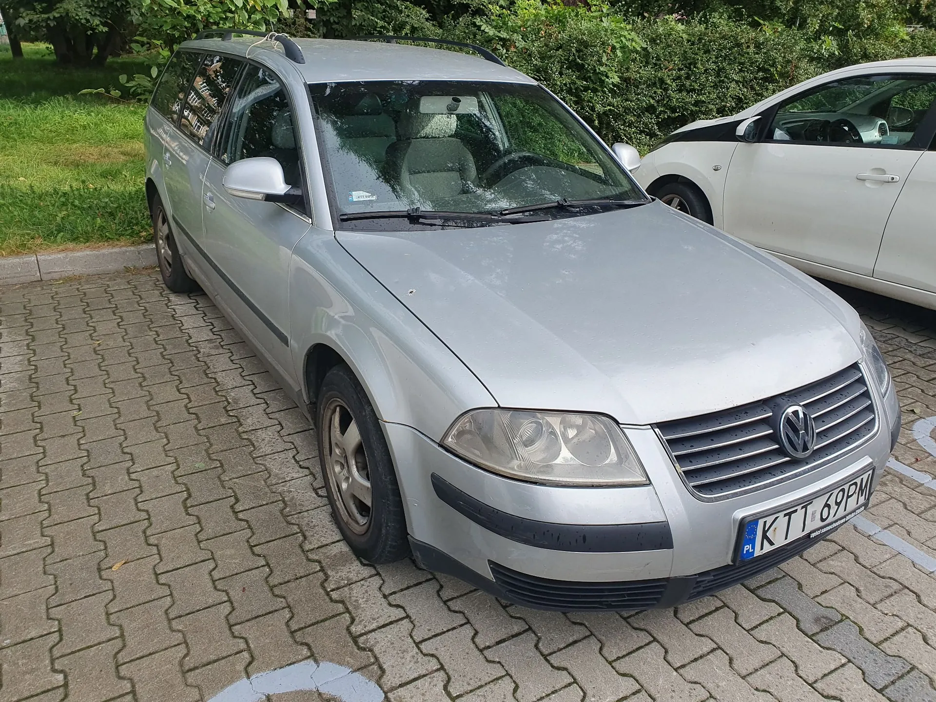 Srebrny samochód osobowy marki Volkswagen w nadwoziu kombi, zaparkowany na kostce brukowej. Pojazd ma polską tablicę rejestracyjną o numerze KTT 69PM. Obok niego stoi biały samochód. W tle widoczna jest zielona trawa i krzewy.