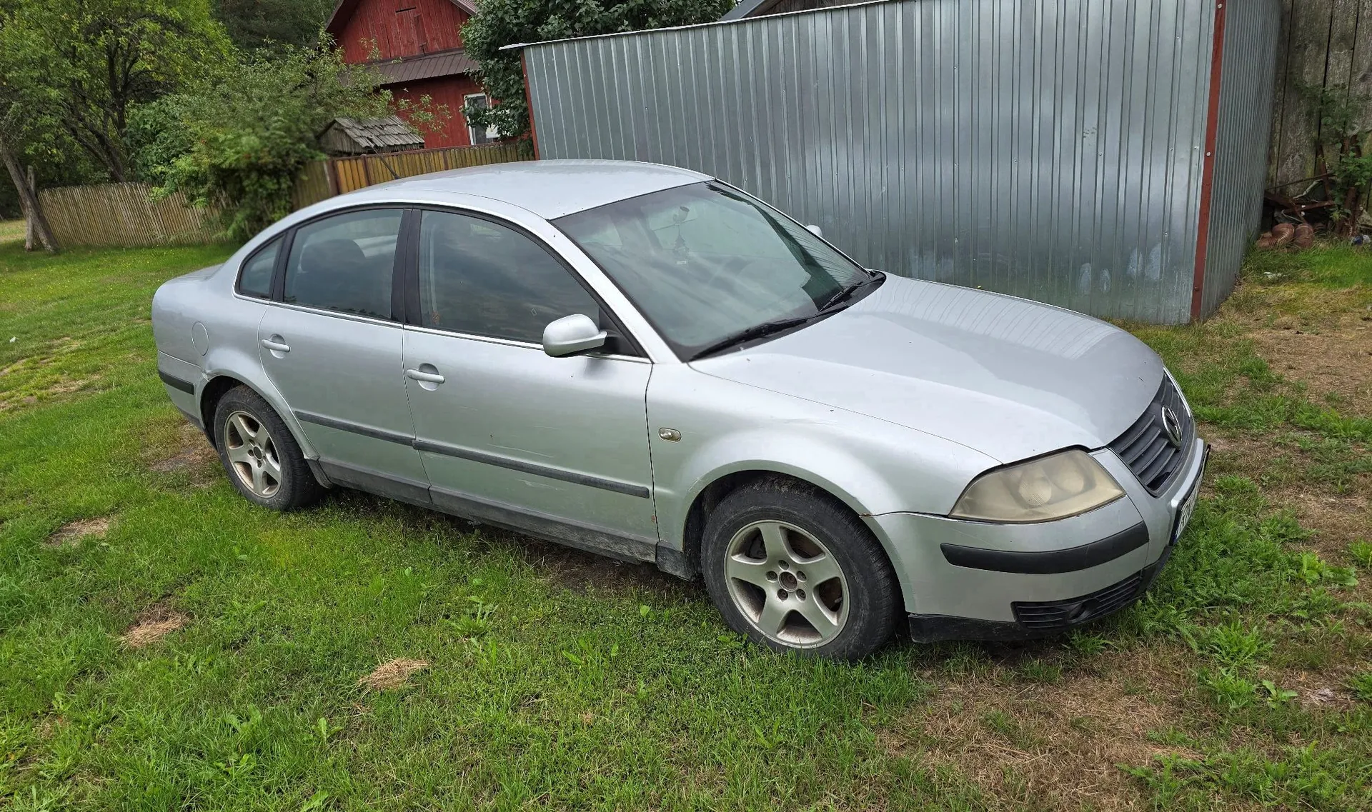 Srebrny samochód osobowy marki Volkswagen Passat zaparkowany na trawiastym terenie. Auto jest ustawione bokiem, przodem w prawą stronę obrazu. W tle widoczny jest szary, blaszany budynek gospodarczy oraz fragment czerwonego drewnianego domu i zielone drzewa.