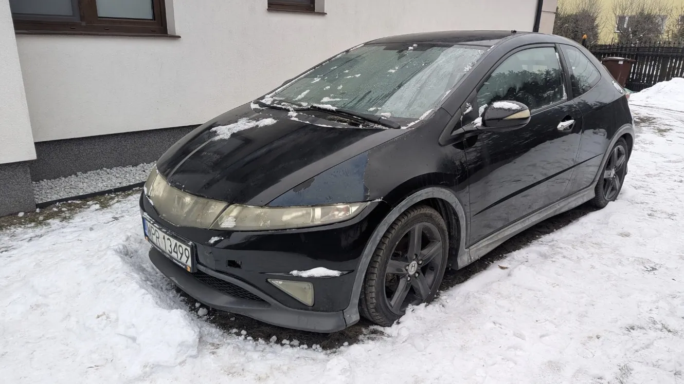 Czarny samochód osobowy typu hatchback zaparkowany na śniegu. Na masce i przedniej szybie widoczne są resztki śniegu. Tablica rejestracyjna z numerem WPR 13499. W tle budynek z białą elewacją, oknem z brązowym obramowaniem oraz ogrodzenie. Podłoże pokryte śniegiem, częściowo odsłonięta ziemia i żwir wokół budynku.