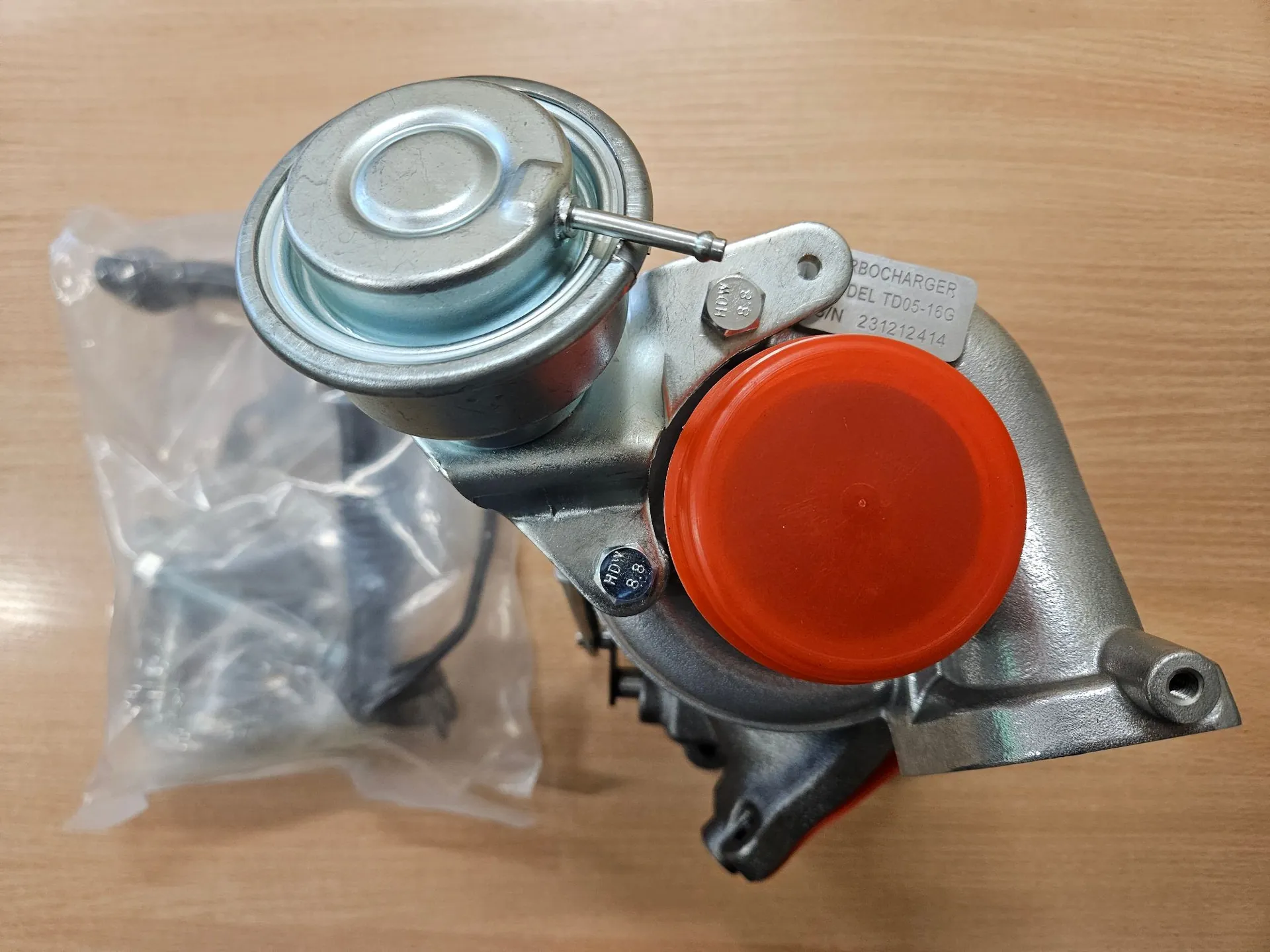 Metalowa turbosprężarka leżąca na drewnianym blacie. Urządzenie posiada srebrny korpus, okrągły element na górze oraz pomarańczową plastikową zaślepkę zamykającą wlot powietrza. Obok turbosprężarki znajduje się przezroczysta foliowa torebka z elementami montażowymi. Na obudowie widoczna jest biała tabliczka znamionowa z tekstem: 'BOCHARGER', 'DEL TD05-16G', 'N 231212414'.