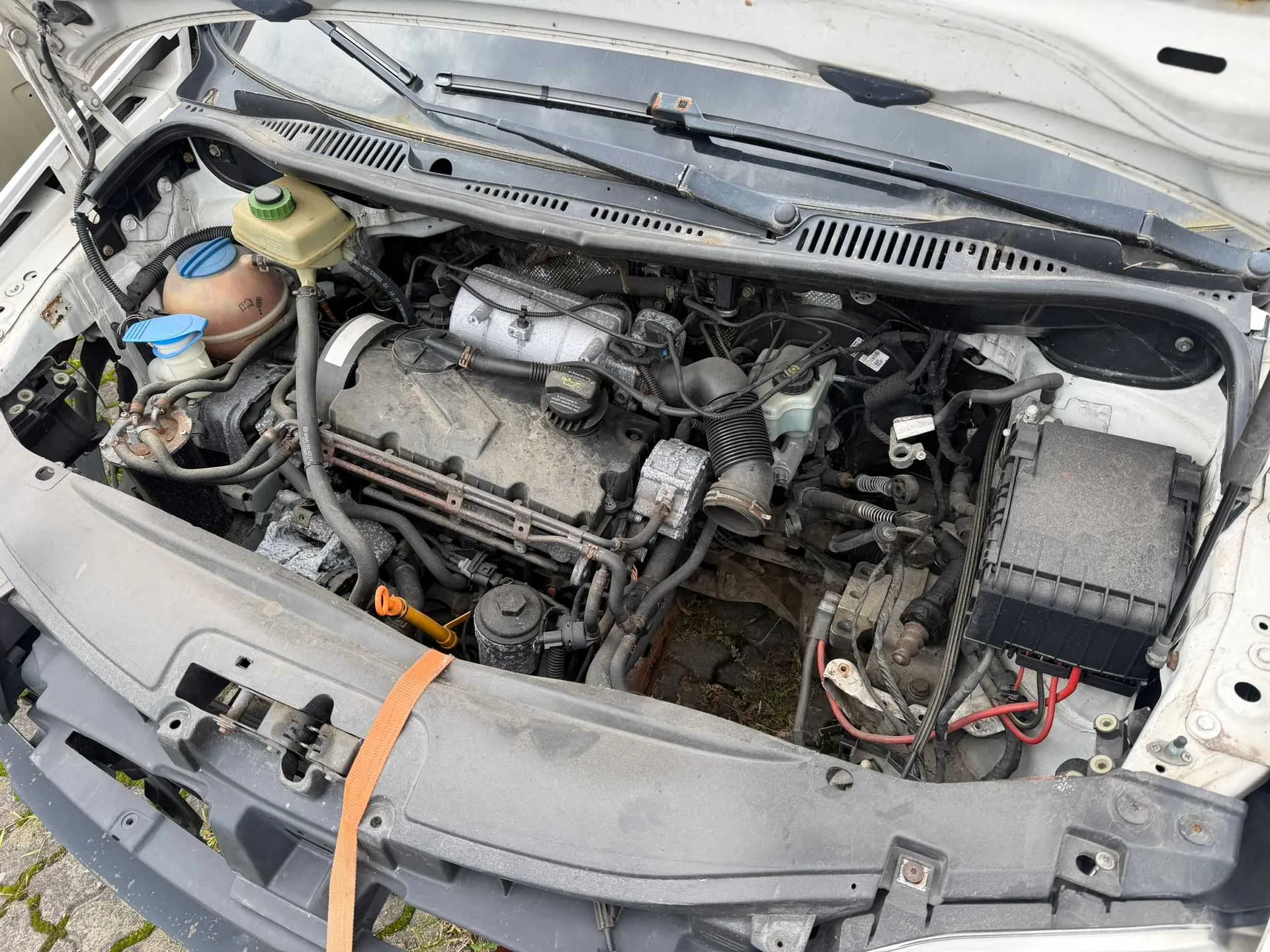 Widok z góry na komorę silnika białego samochodu z otwartą maską. Widoczny jest silnik spalinowy z różnymi przewodami, wężami i elementami mechanicznymi, a także zbiorniki z płynami eksploatacyjnymi po lewej stronie oraz czarna skrzynka bezpieczników po prawej. W dolnej części zdjęcia widoczny jest fragment przedniego zderzaka i pas przedniego, przez który przechodzi pomarańczowy pas transportowy.