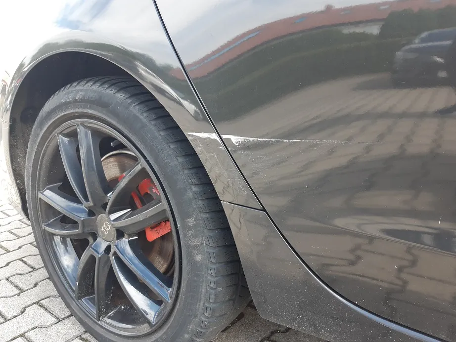 Zbliżenie na przednie lewe koło czarnego samochodu. Widoczna jest czarna felga z logo Audi oraz opona. Na nadkolu, nad oponą, znajduje się pozioma, biała rysa w lakierze.