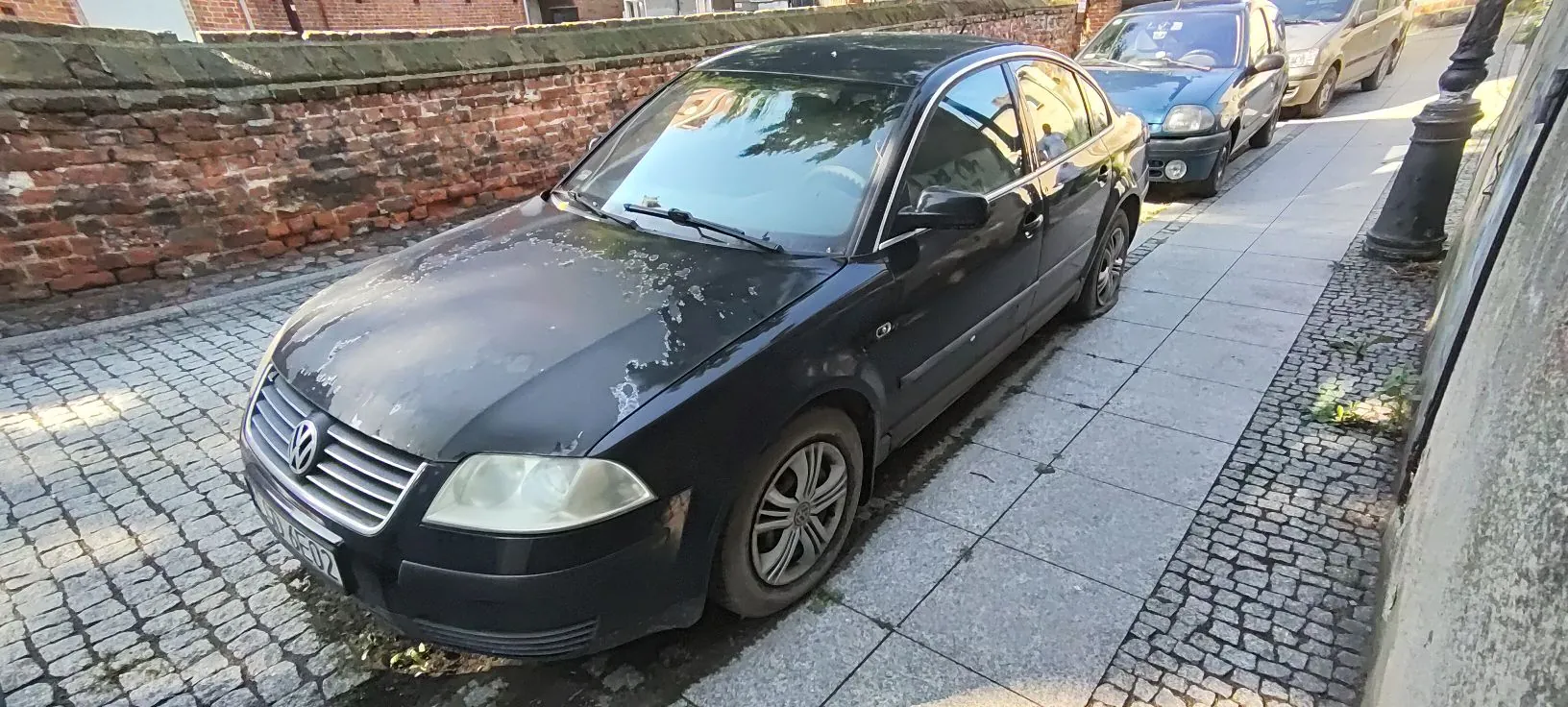 Czarny samochód osobowy marki Volkswagen zaparkowany na chodniku z płyt betonowych i kostki brukowej, obok starego ceglanego muru. W tle widoczne są inne zaparkowane samochody oraz fragment budynku.