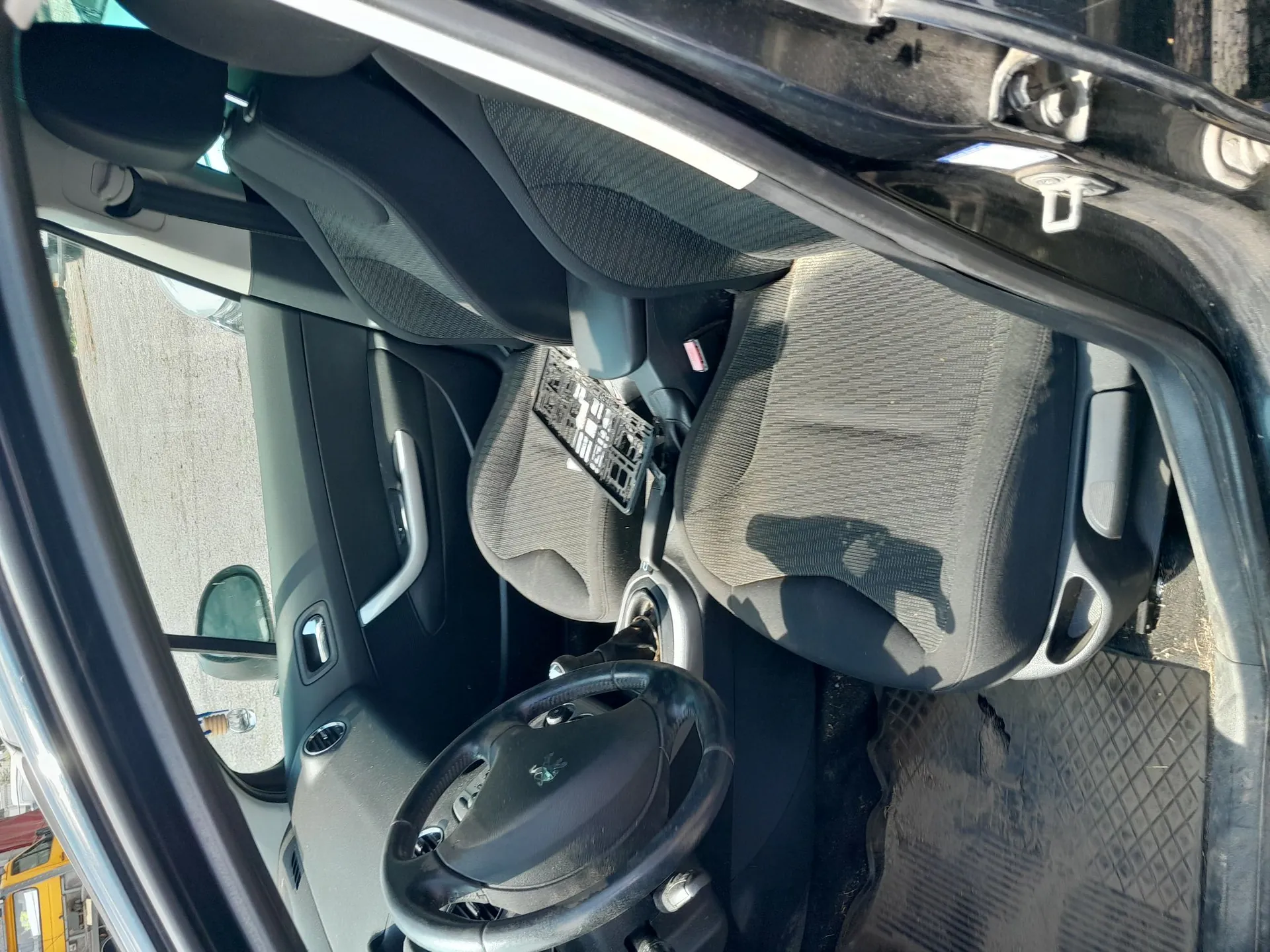 Wnętrze samochodu widziane z otwartych drzwi kierowcy. Widoczna jest czarna kierownica z logo marki Peugeot, ciemnoszare fotele z materiałowej tapicerki oraz elementy deski rozdzielczej i boczny panel drzwi. Na oparciu fotela pasażera znajduje się przymocowana czarna tabliczka z tekstem i tabelami.