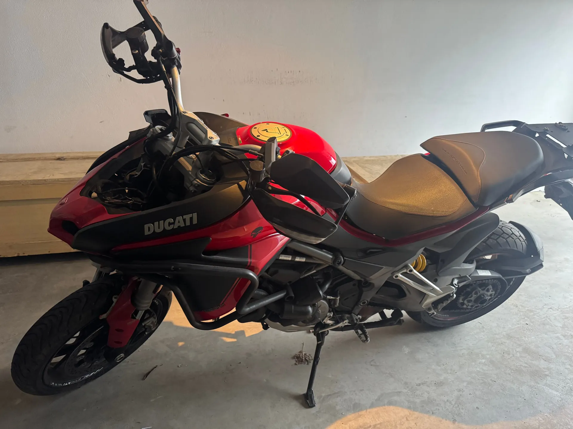 Czerwono-czarny motocykl marki Ducati zaparkowany w pomieszczeniu na białym tle. Motocykl stoi na bocznej stopce, widoczny jest napis 'DUCATI' na boku owiewki. Pojazd posiada czarną kanapę, czerwony zbiornik paliwa oraz czarne osłony dłoni na kierownicy.