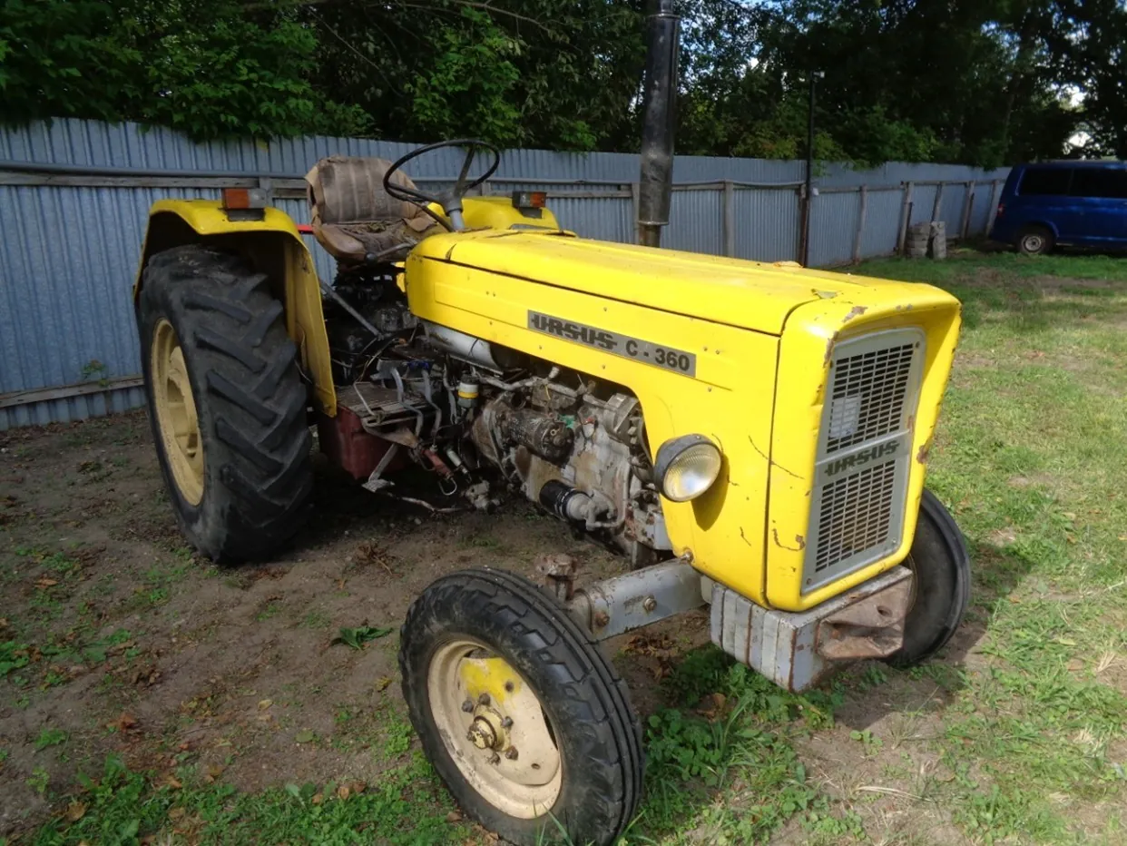 Żółty traktor marki Ursus, model C-360, zaparkowany na trawiasto-piaszczystym podłożu. Na masce widnieje napis 'URSUS C-360'. Maszyna ma duże tylne koła z bieżnikiem i mniejsze przednie koła. W tle znajduje się szary, falisty płot z blachy oraz zielone drzewa. Po prawej stronie widoczny jest fragment niebieskiego samochodu.