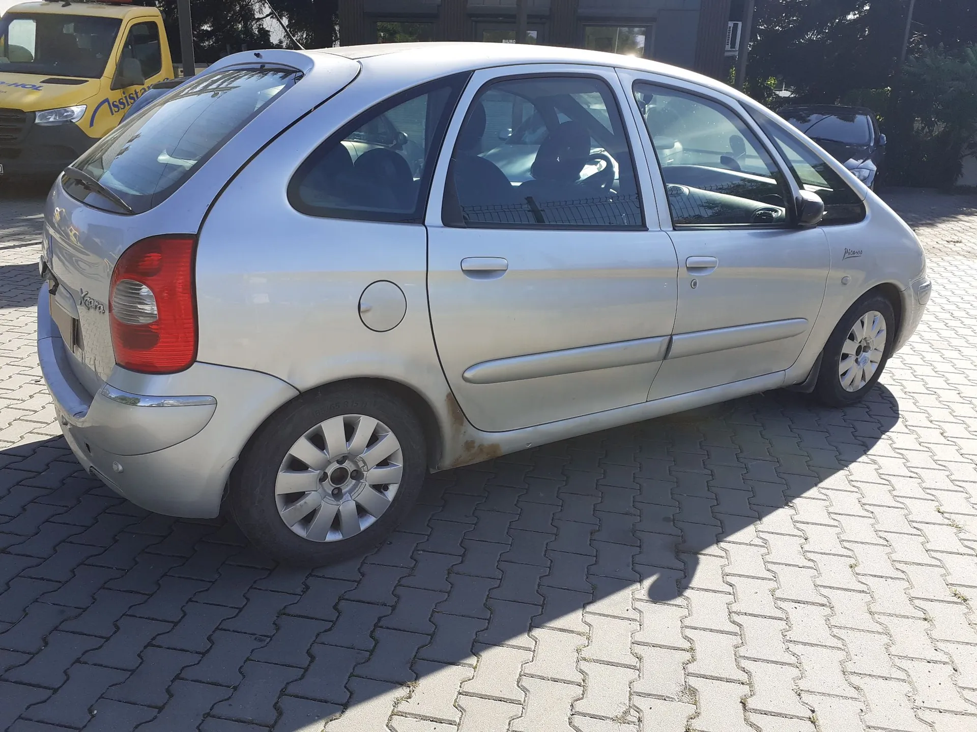 Srebrny samochód osobowy marki Renault Scenic zaparkowany na kostce brukowej. Pojazd jest widoczny z boku i z tyłu, z widoczną korozją na dolnej krawędzi drzwi kierowcy. W tle widoczny jest żółty samochód dostawczy z napisem 'Assis'.