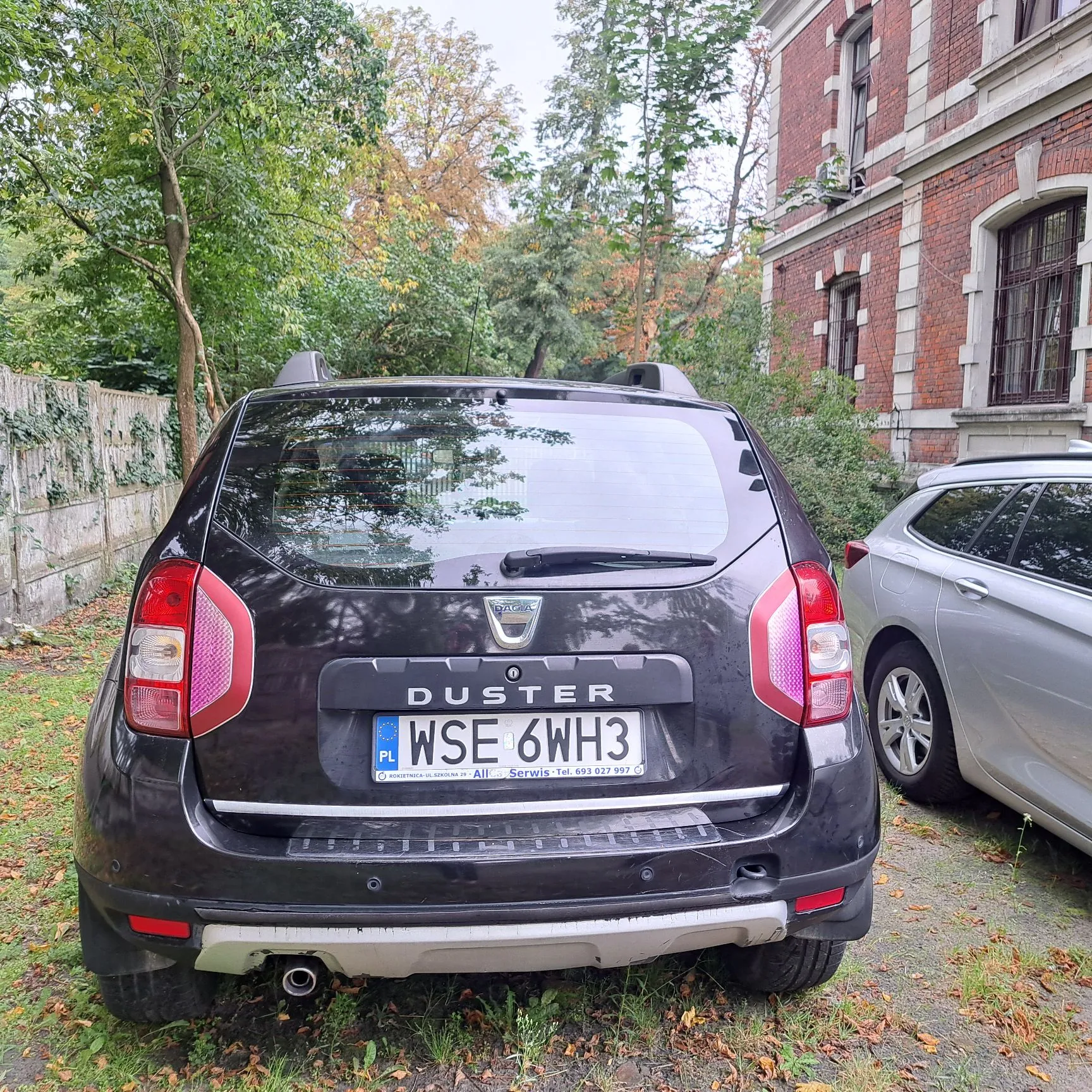 Tylny widok czarnego samochodu marki Dacia Duster zaparkowanego na trawiastym podłożu. Na klapie bagażnika widnieje napis DUSTER oraz polska tablica rejestracyjna o numerze WSE 6WH3. Poniżej tablicy znajduje się naklejka z napisem 'All Serwis Tel. 693 627 997'. Po prawej stronie widoczny jest fragment srebrnego samochodu, a w tle ceglany budynek oraz zielone drzewa.