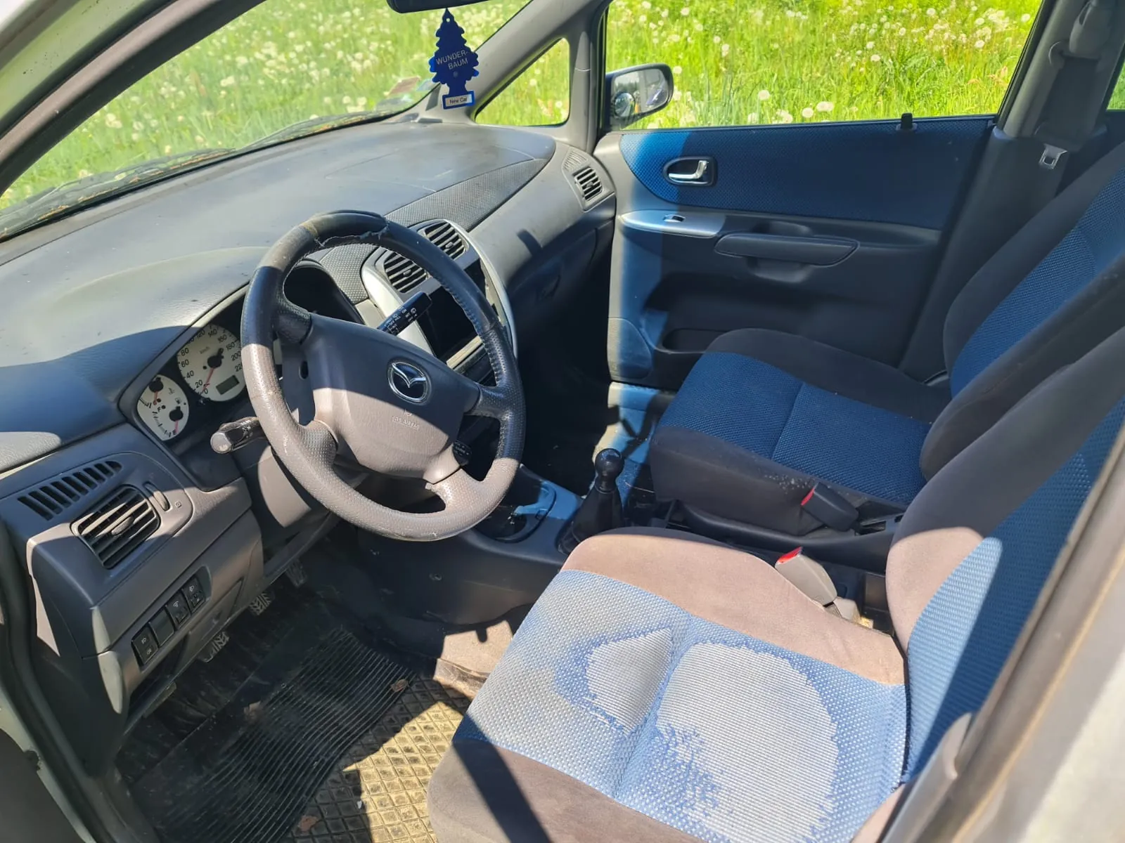 Wnętrze samochodu marki Mazda z widocznym kierownicą z logo, tablicą przyrządów, niebiesko-szarymi siedzeniami i otwartymi drzwiami. Na lusterku wstecznym wiszący zapach samochodowy z napisem "WUNDER-BAUM". Za oknem zielona trawa z białymi kwiatami. Deska rozdzielcza i elementy wnętrza są w kolorze szarym, a podłoga pokryta czarną wykładziną.