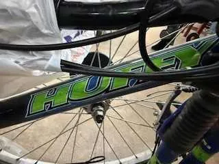 Zbliżenie na rame roweru z widocznym napisem 'HUFFY' w kolorach zielonym i niebieskim. Widoczne są również części roweru: koło przednie z promieniami, przewody hamulcowe oraz tło z betonowej powierzchni.